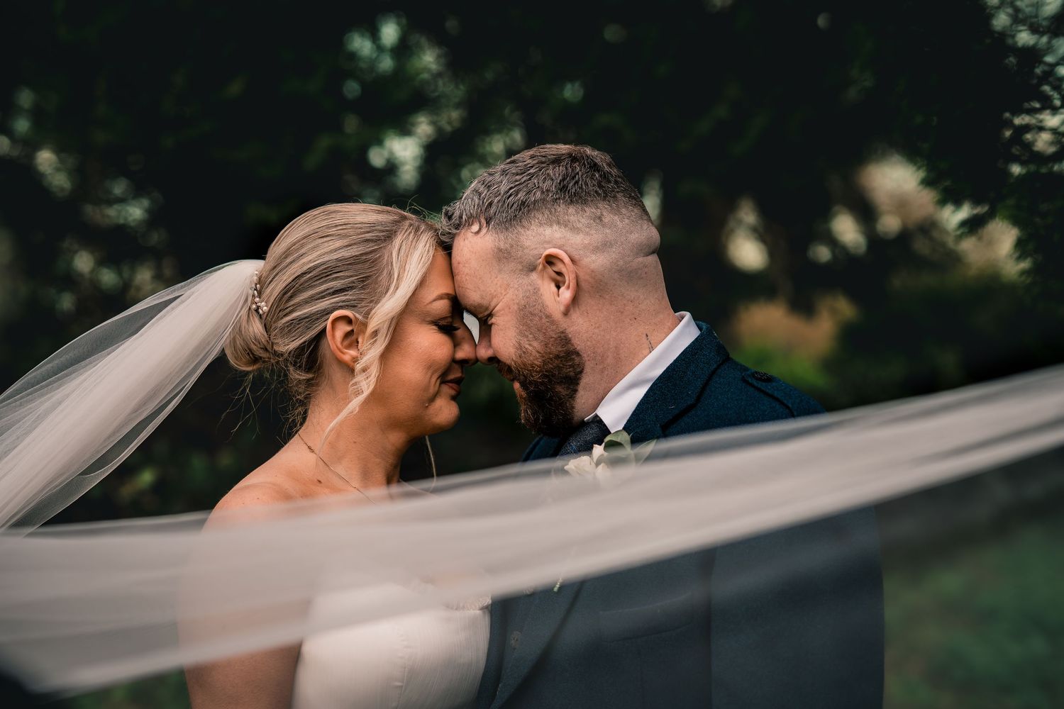 A bride and groom share an intimate forehead touch moment, with her veil flowing in the breeze at their outdoor wedding.