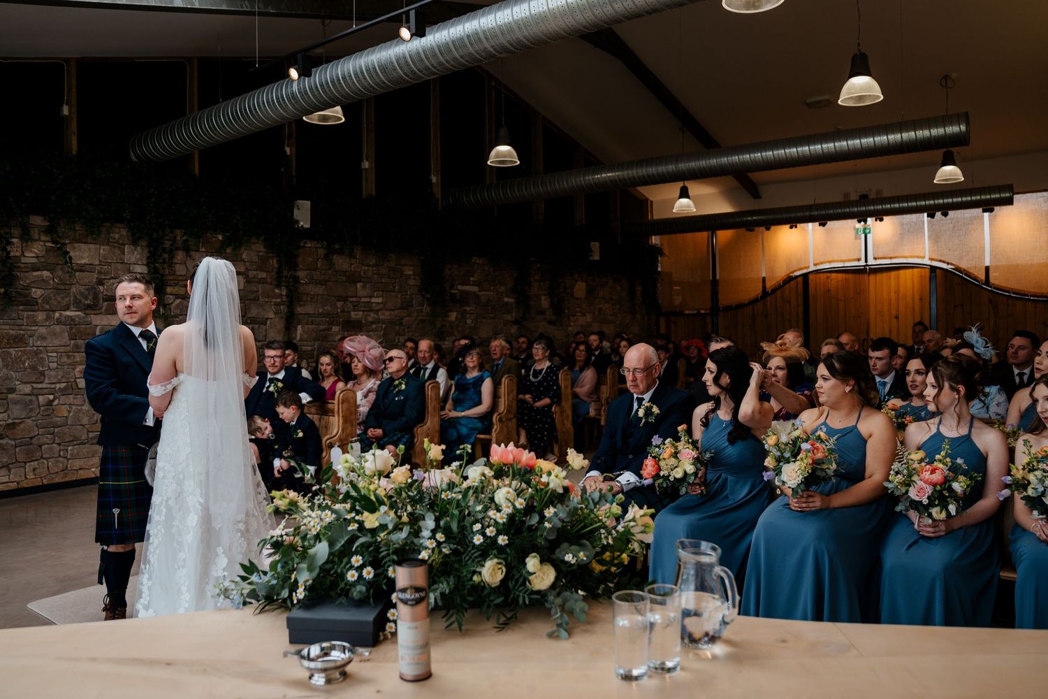 Bride and groom stand at altar during wedding ceremony with bridesmaids in blue dresses and guests seated in rustic venue.