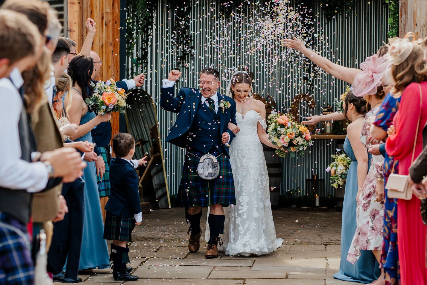Newlywed couple walks through confetti shower, groom in blue tartan kilt, bride in lace gown, guests celebrating outdoors.