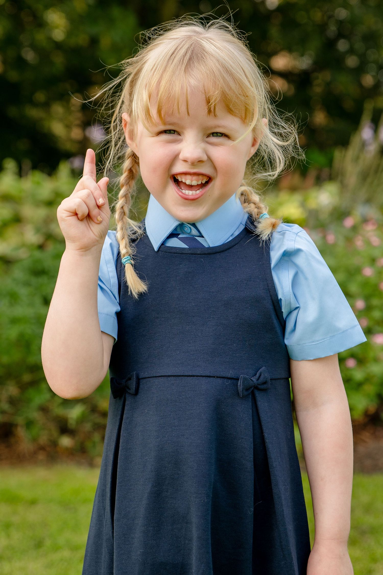 Young girl in school uniform with braids raises index finger, smiling outdoors in a garden setting.