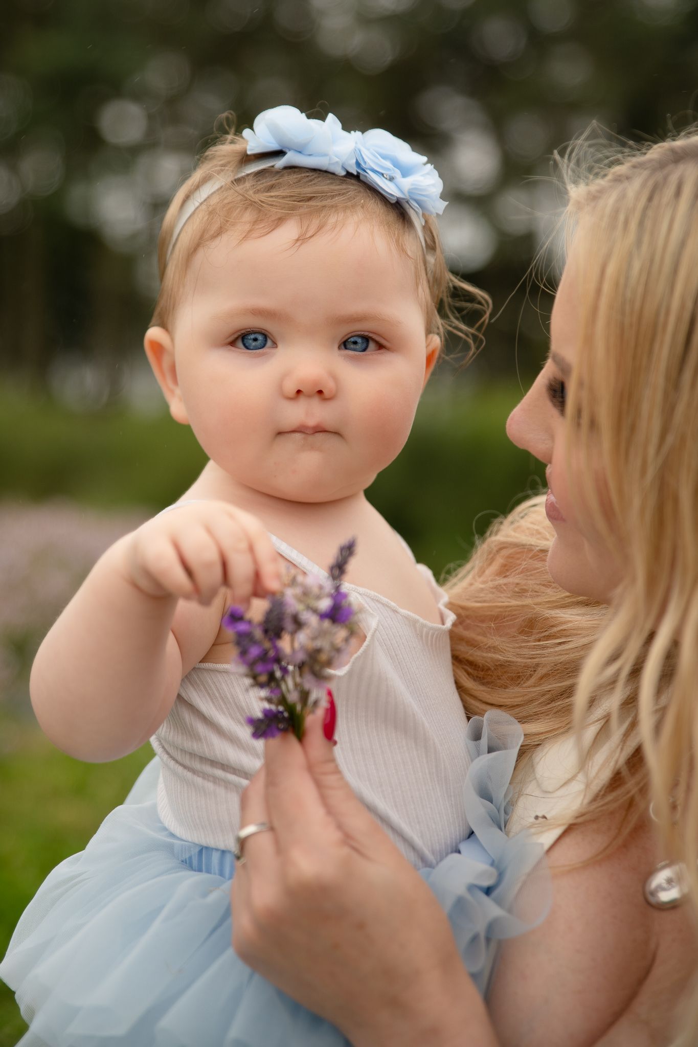 A blue-eyed baby girl in a blue tutu and floral headband holds purple flowers while being held by a blonde woman outdoors.