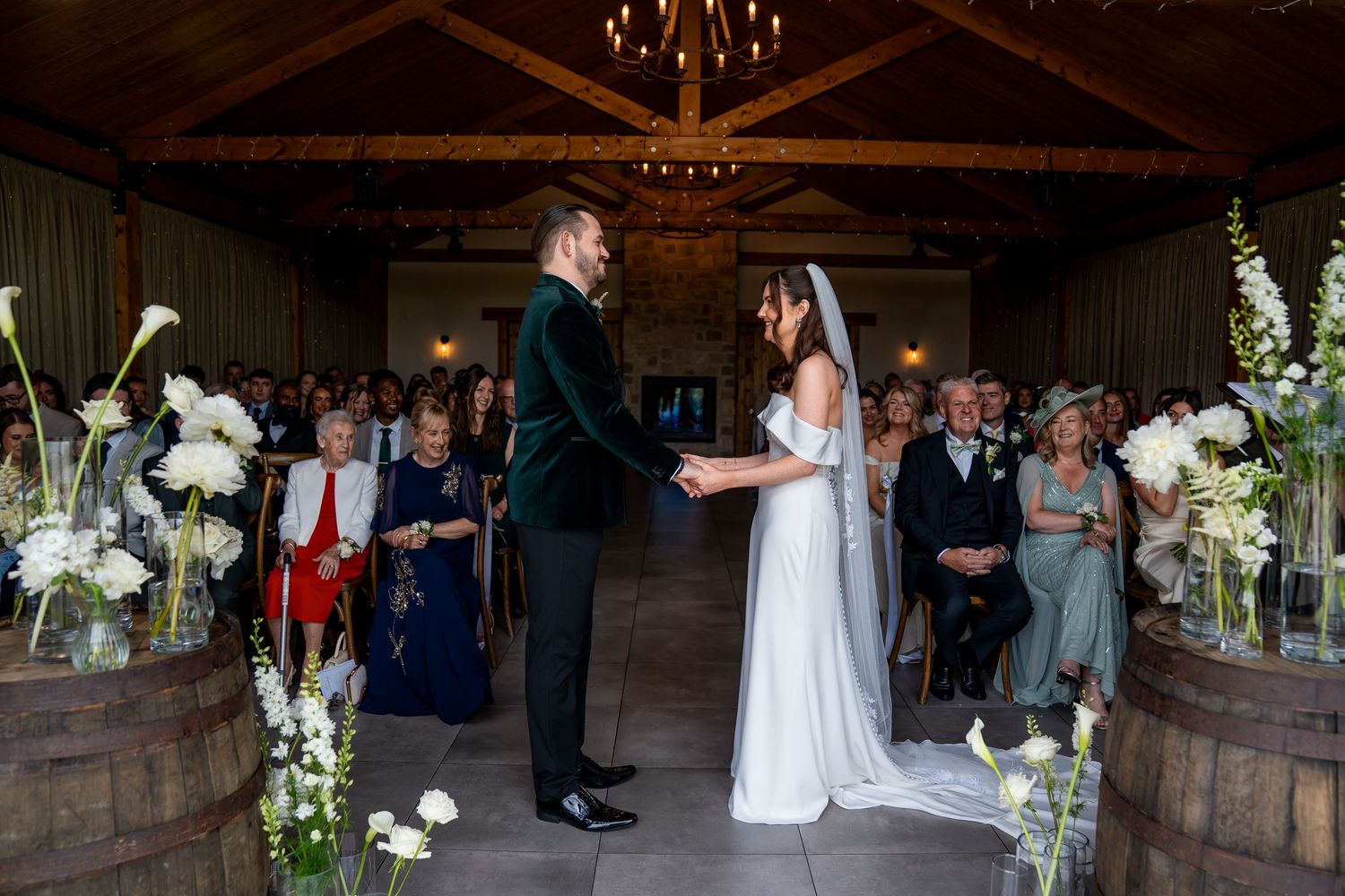 Bride and groom exchange vows in elegant barn wedding venue with white floral arrangements and seated guests.