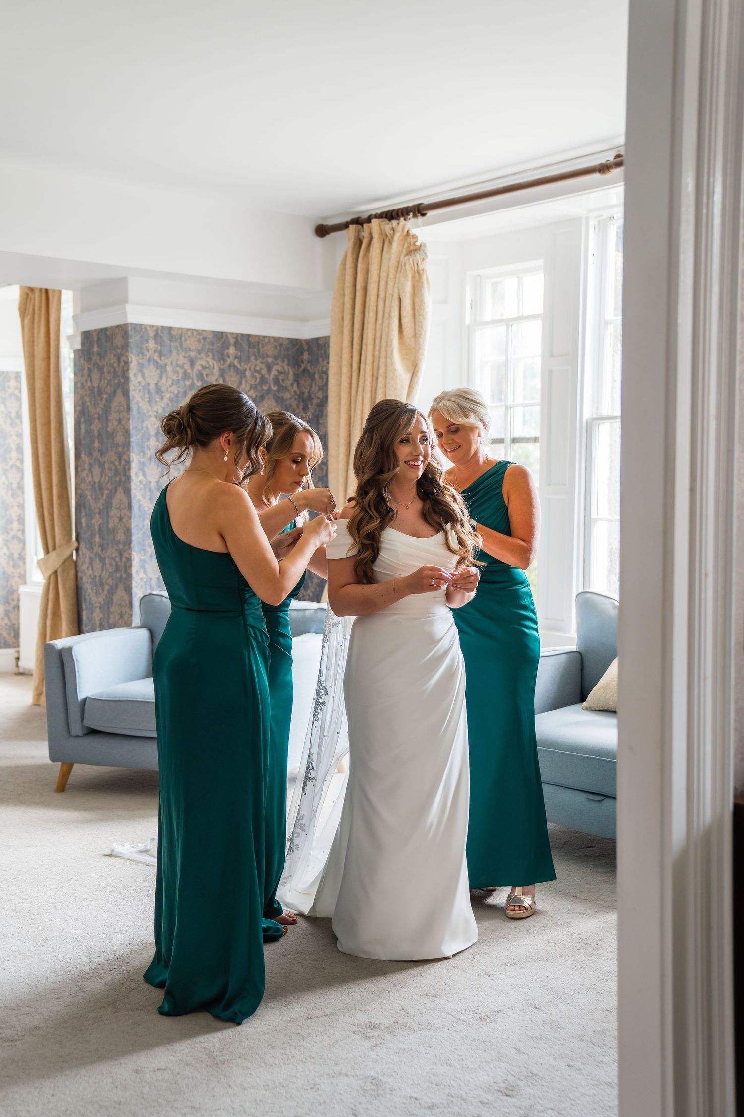 Bride in white gown with two bridesmaids in emerald green dresses helping her prepare in an elegant room.