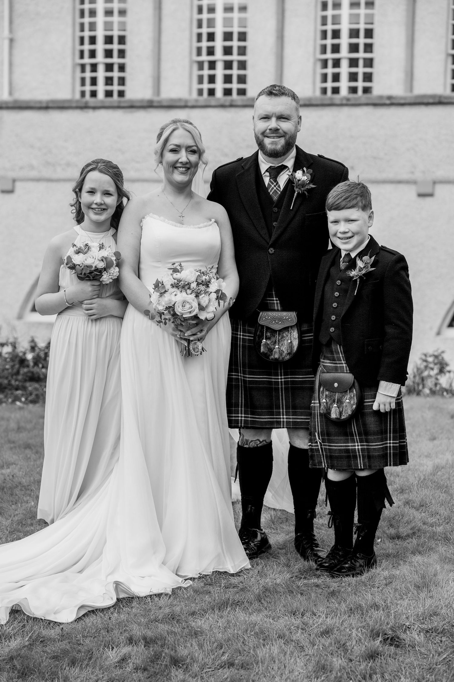 Black and white wedding photo of a bride and groom in Scottish kilt posing with two children outside a historic building.