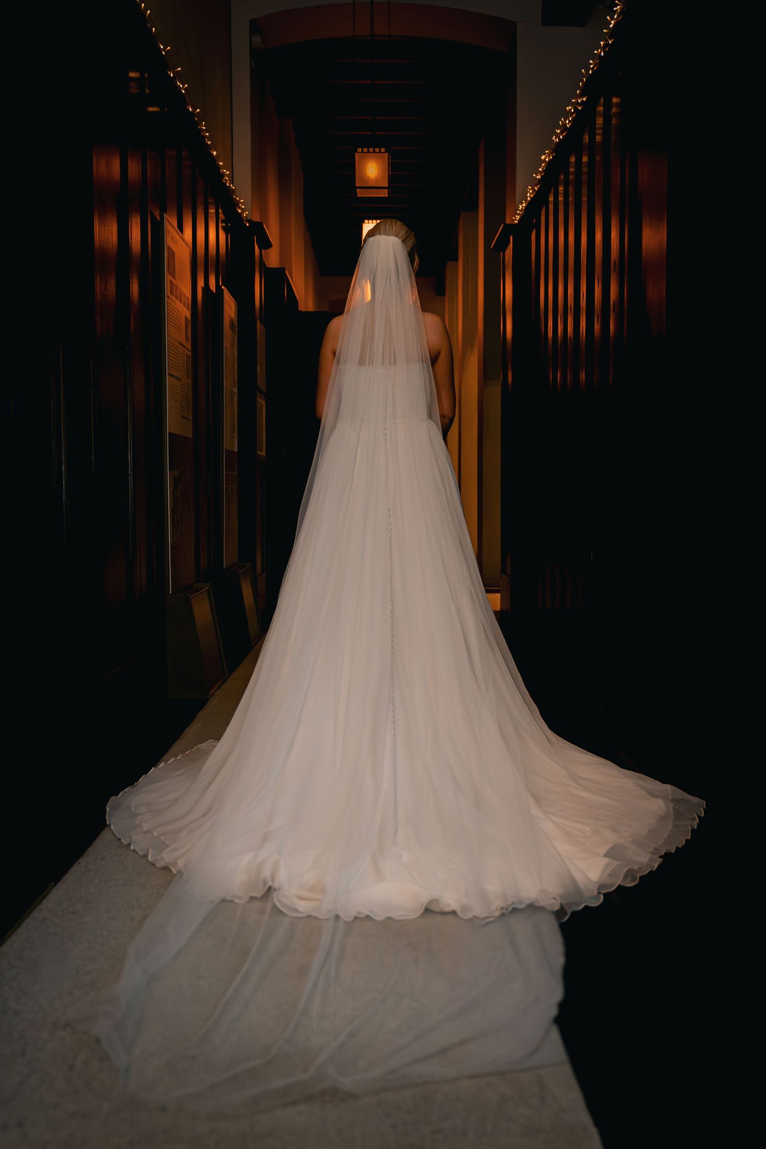 Bride in elegant white wedding gown with long veil and train walks down dimly lit corridor with warm fairy lights.