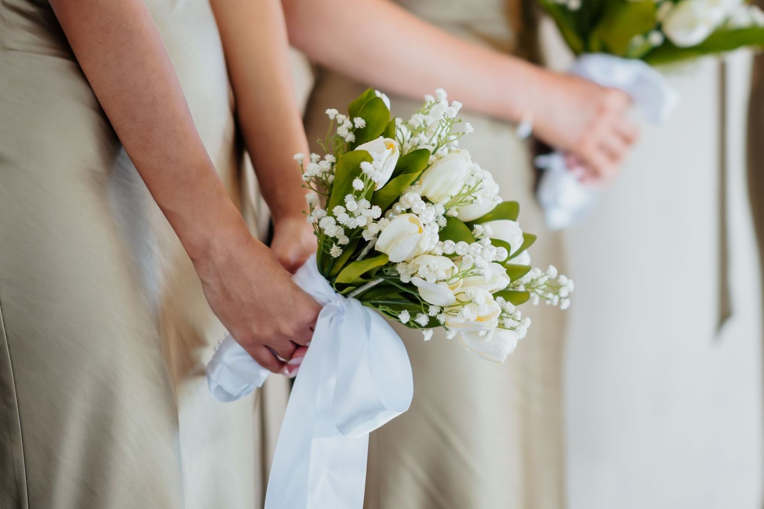 A delicate white lily of the valley bouquet wrapped in white ribbon held against a beige dress.