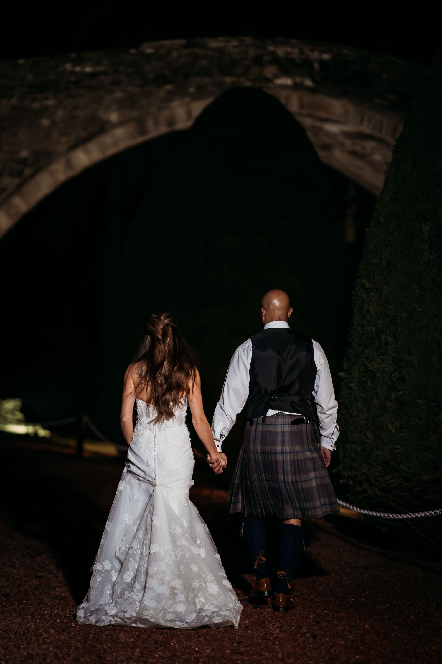 Newlywed couple holding hands walking into dark stone tunnel, with groom wearing traditional Scottish kilt.