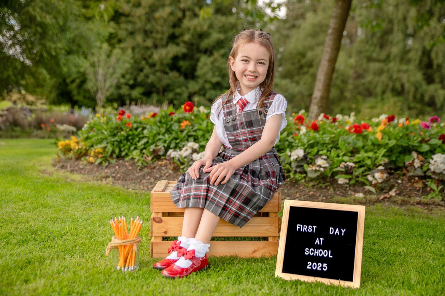 Girl in plaid school uniform sitting on wooden crate with pencils and First Day of School 2023 sign.