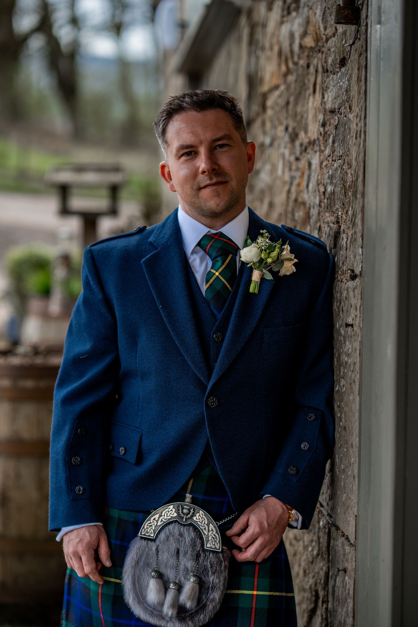 Groom in blue Scottish kilt outfit with tartan sporran and floral boutonniere leaning against stone wall.