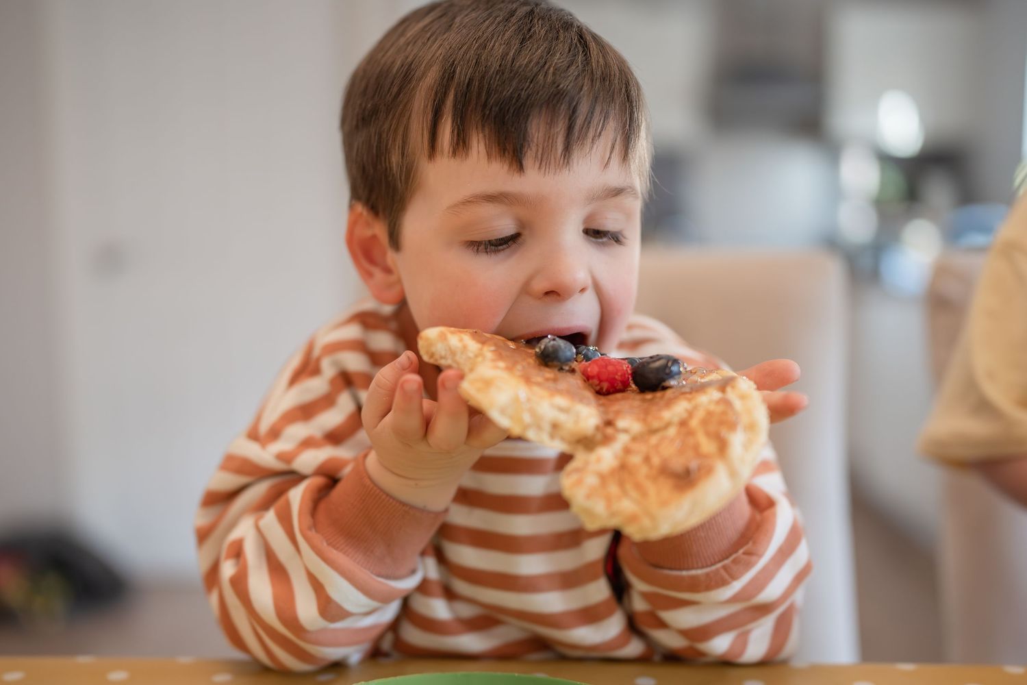 Young boy in striped shirt eating a slice of toast topped with fresh blueberries and raspberries.