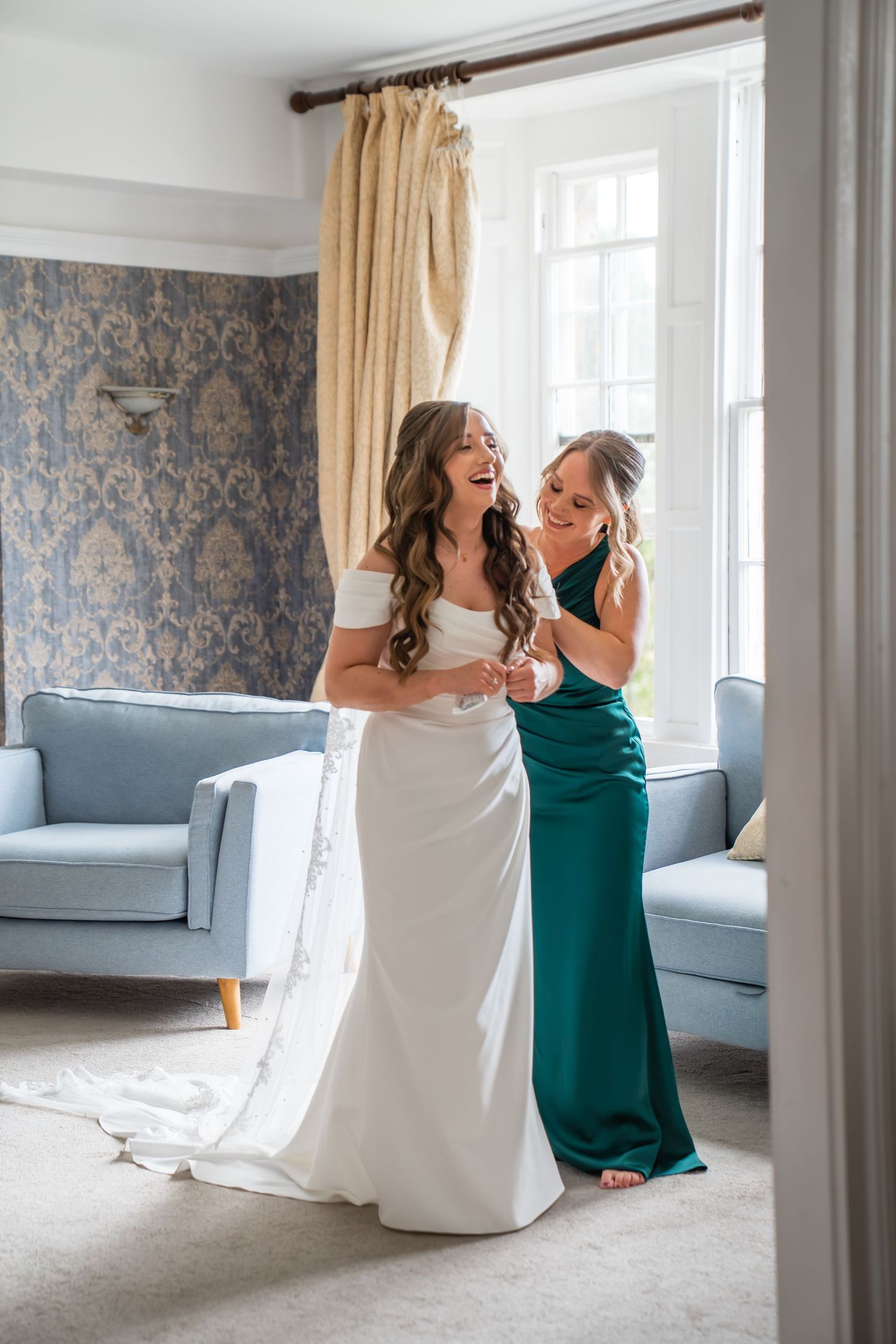 Bride in white gown laughs with bridesmaid in emerald dress while getting ready in elegant blue room.