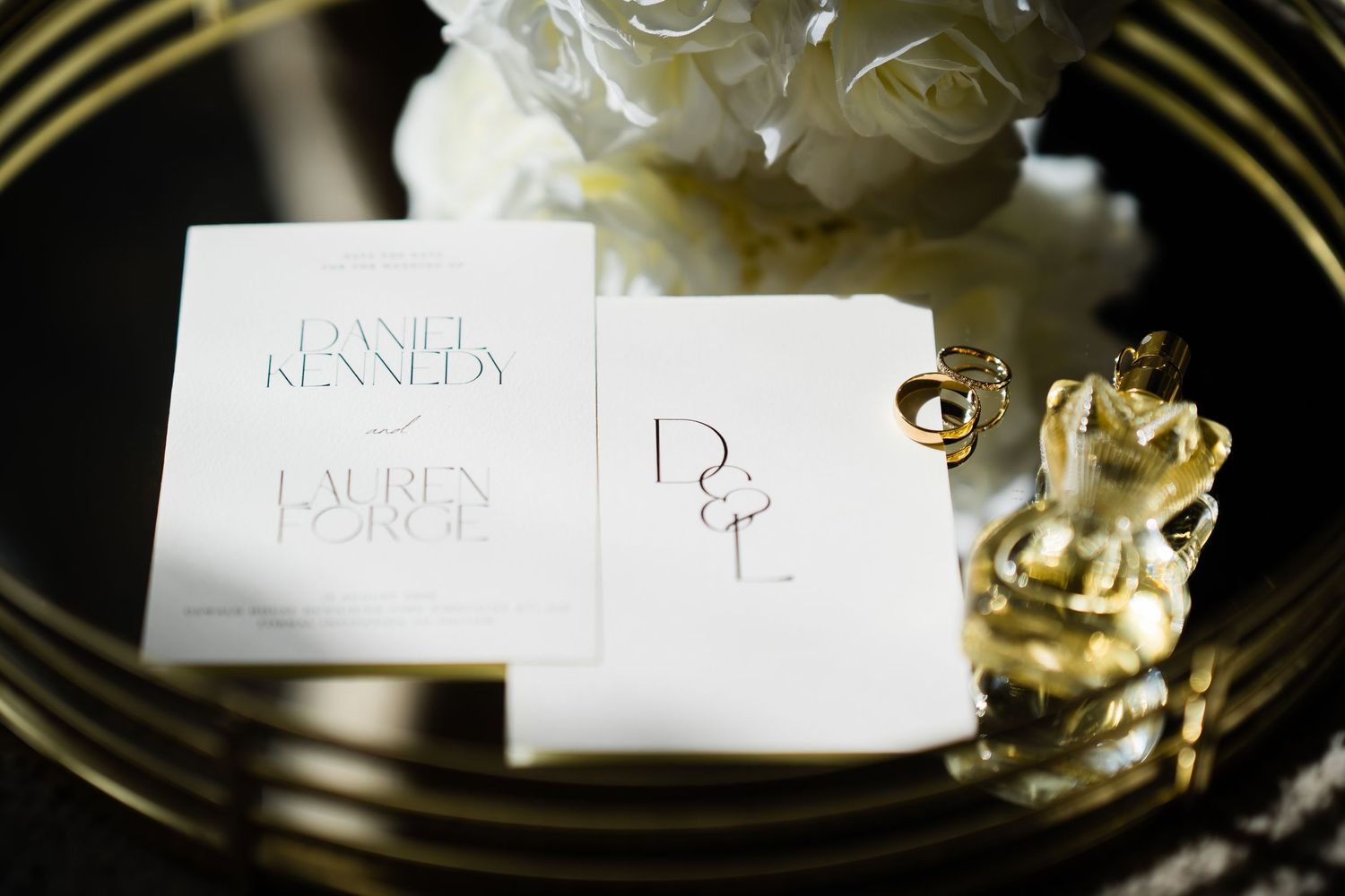 Wedding stationery with Daniel Kennedy and Lauren Force names, gold rings, and white floral arrangement on mirrored tray.
