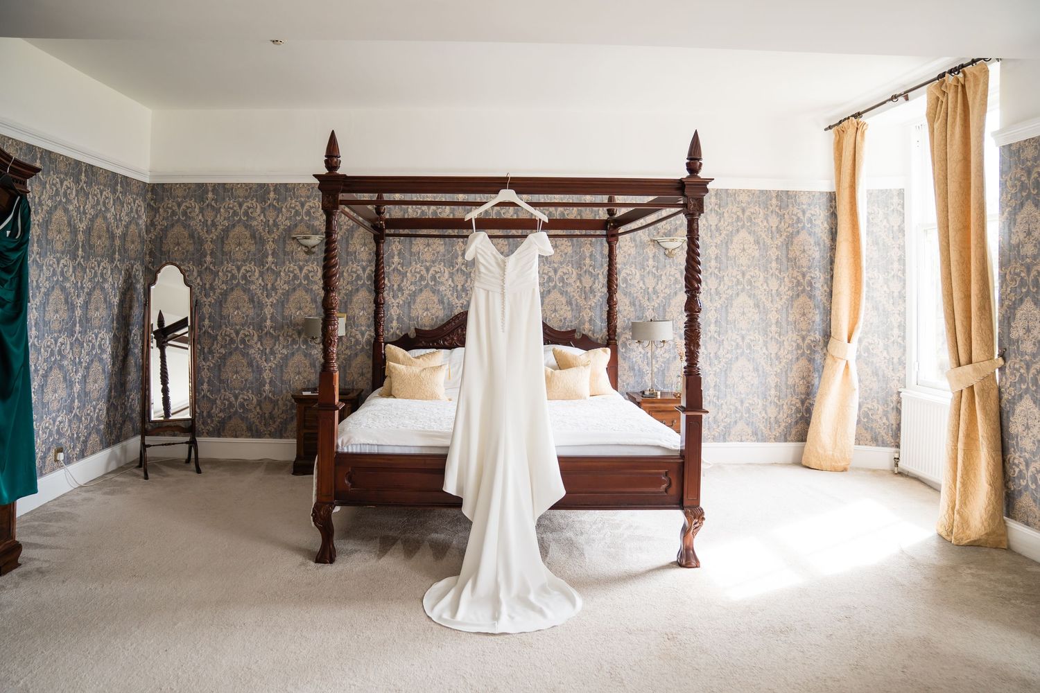 White wedding dress hanging on a four-poster bed in an elegant room with damask wallpaper and gold curtains.