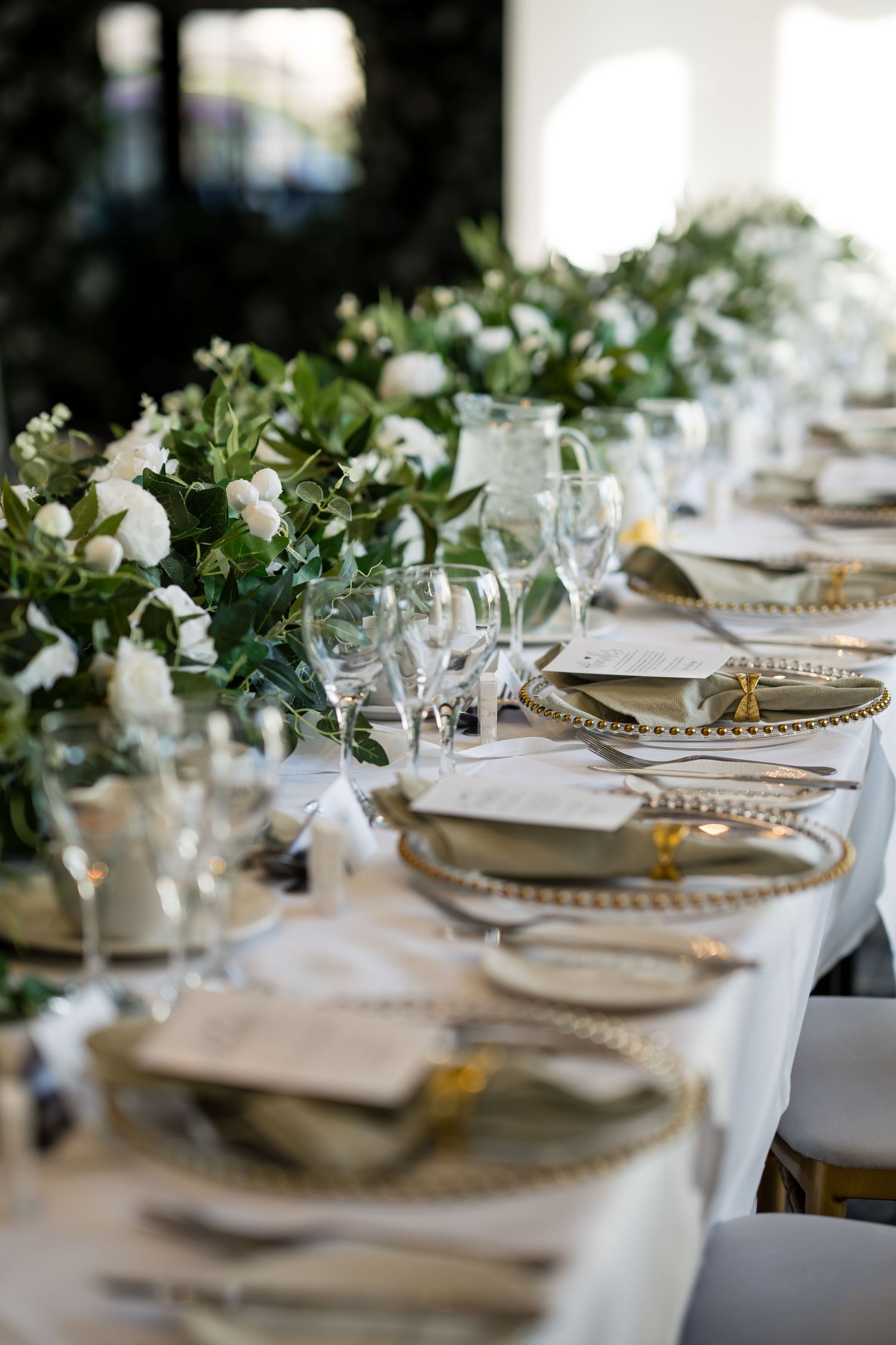 Elegant wedding reception table with gold charger plates, crystal glasses, and white floral greenery centerpieces on white linen.
