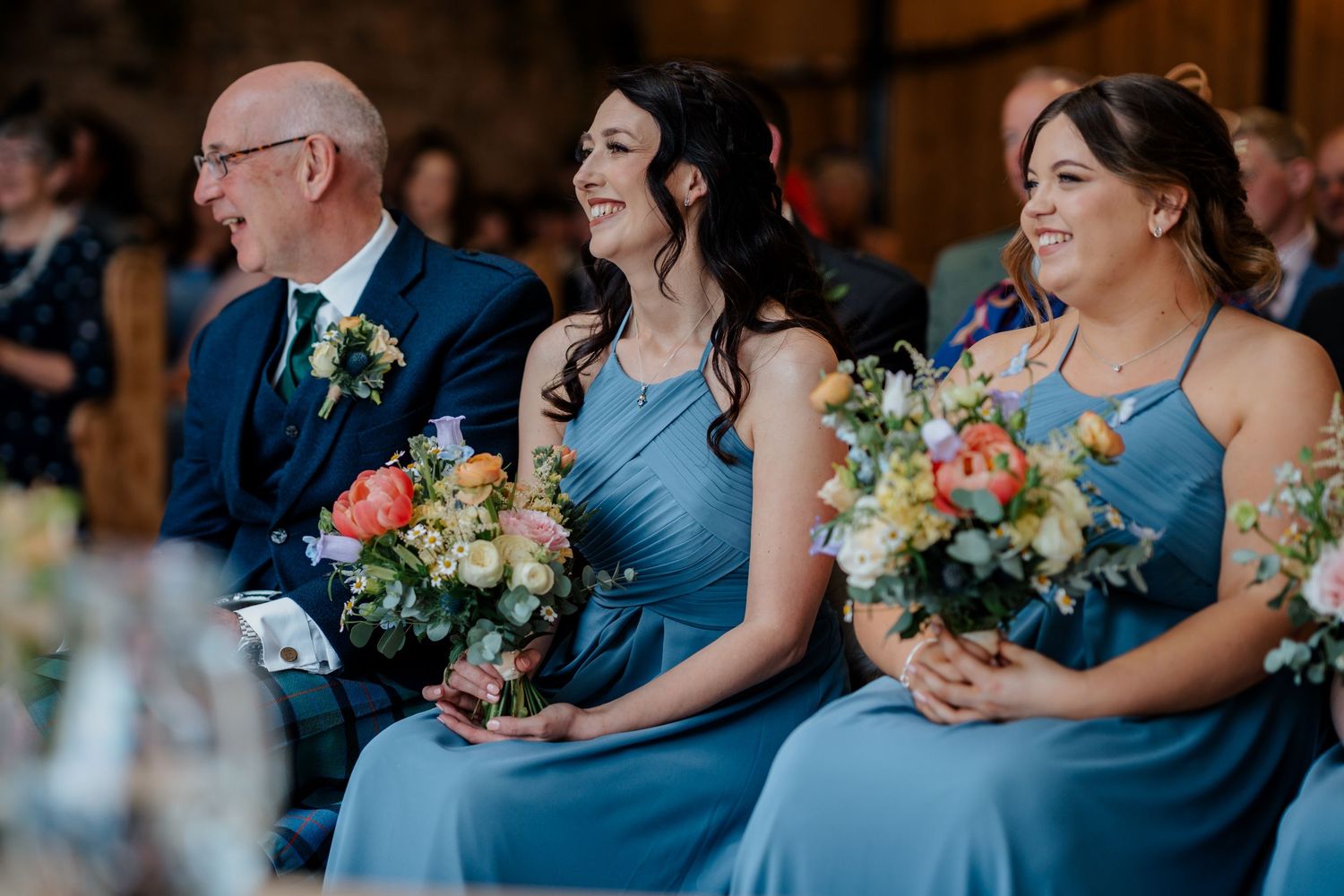 Two bridesmaids in blue dresses holding colorful bouquets smile during a wedding ceremony indoors.