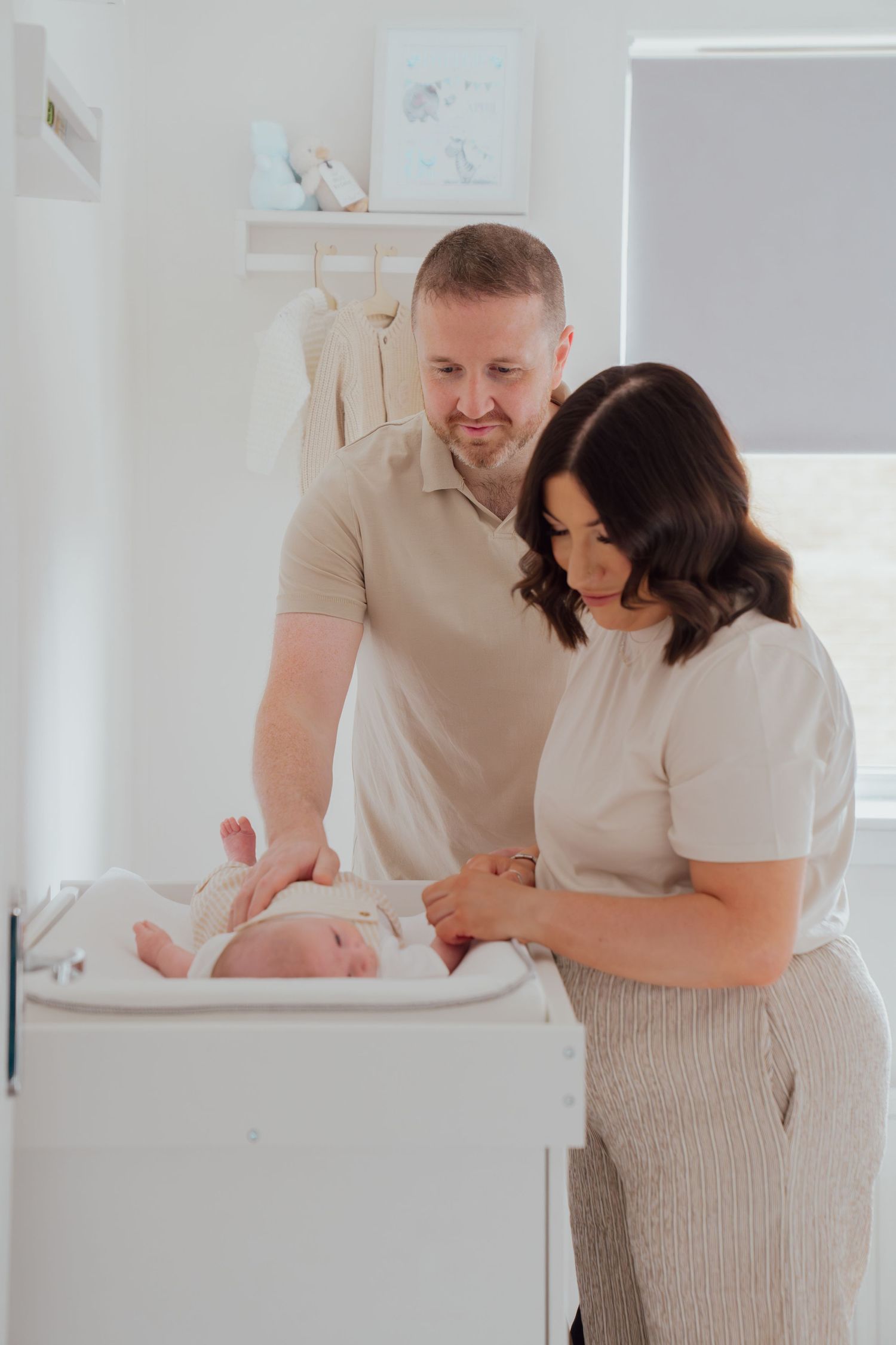 New parents lovingly tend to their newborn baby on a changing table in a bright white nursery room.