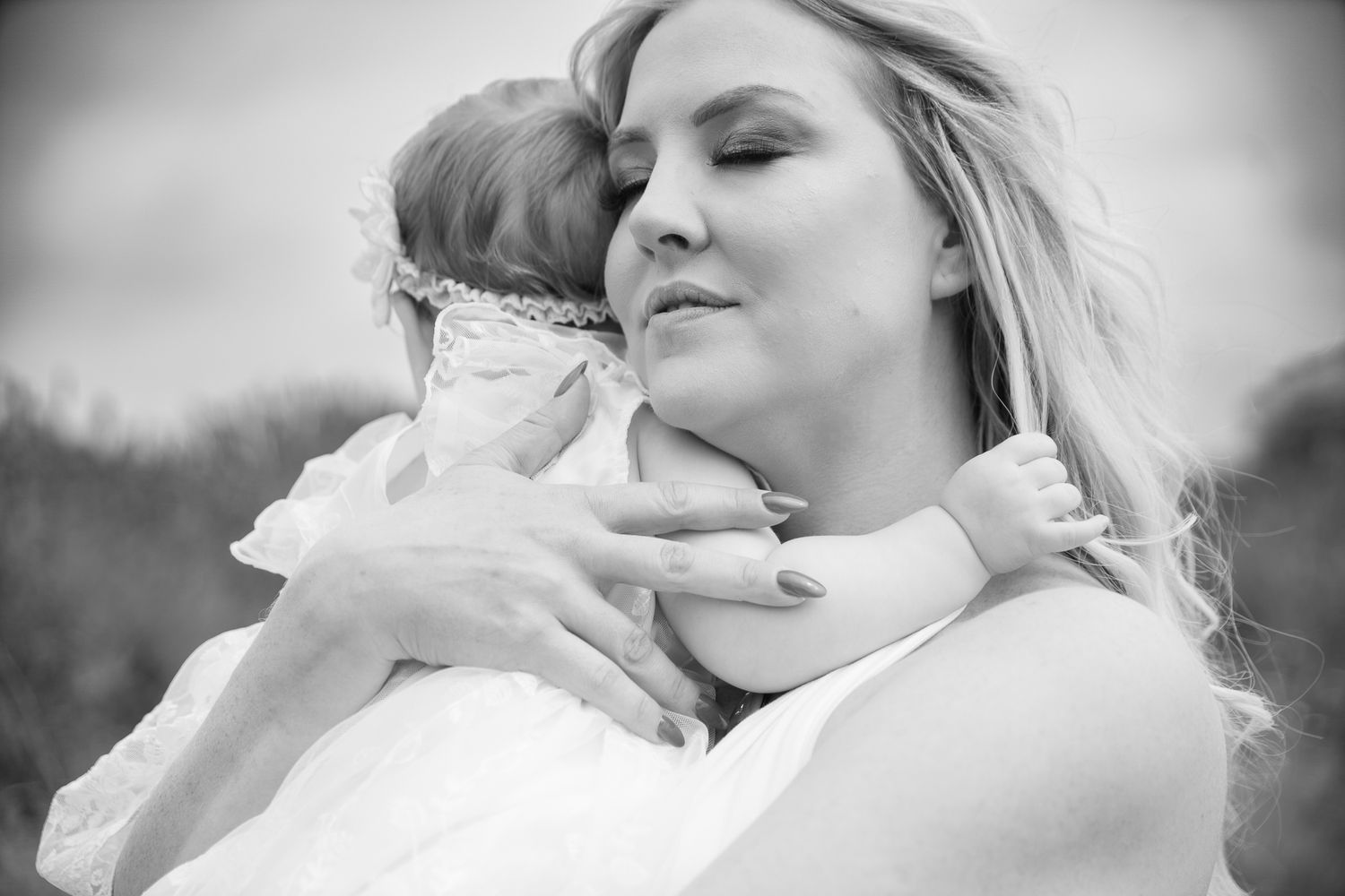 Black and white photo of a blonde woman tenderly embracing a baby wearing a floral headband outdoors.