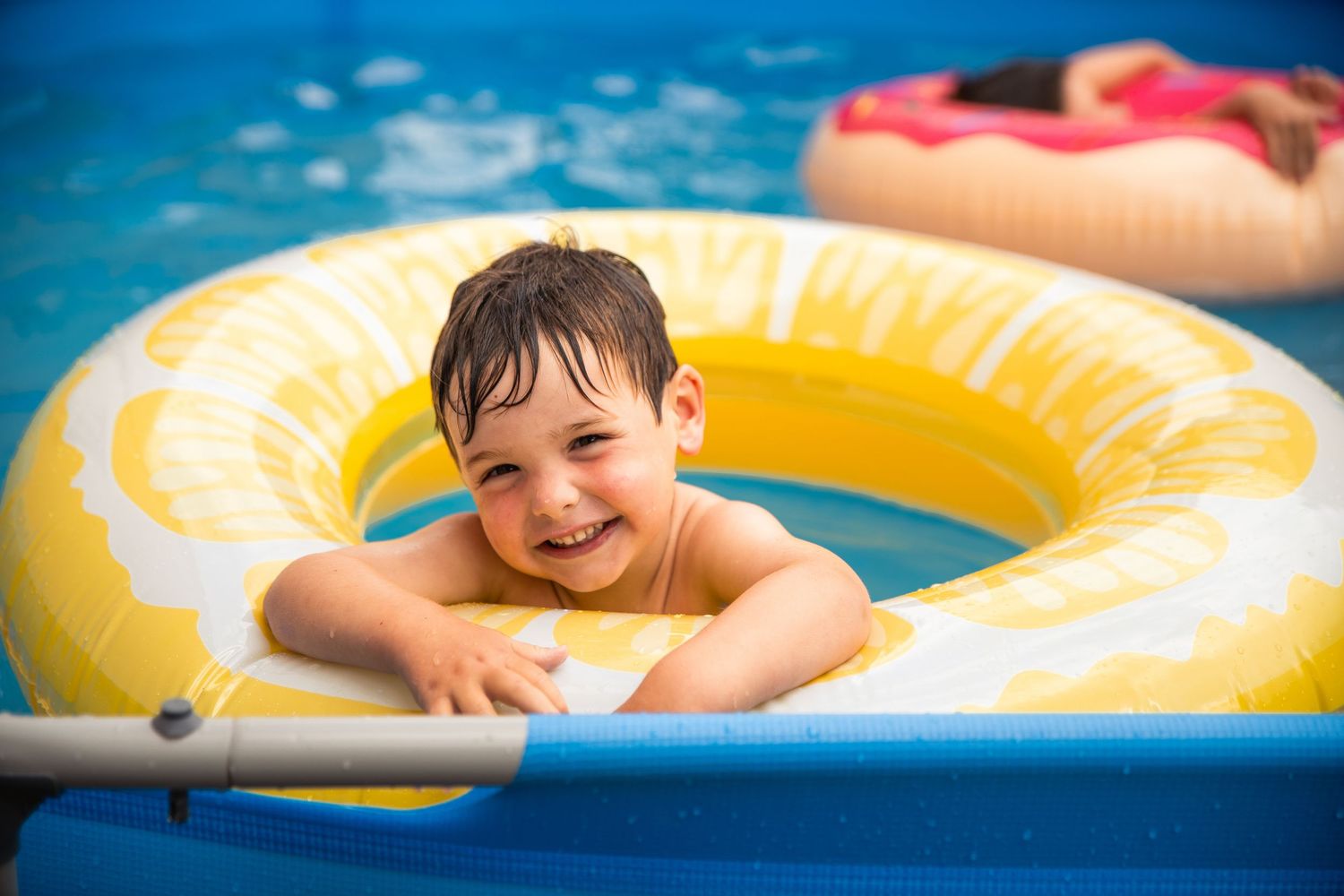 Smiling toddler boy playing in yellow inflatable ring float in a swimming pool on a sunny day.