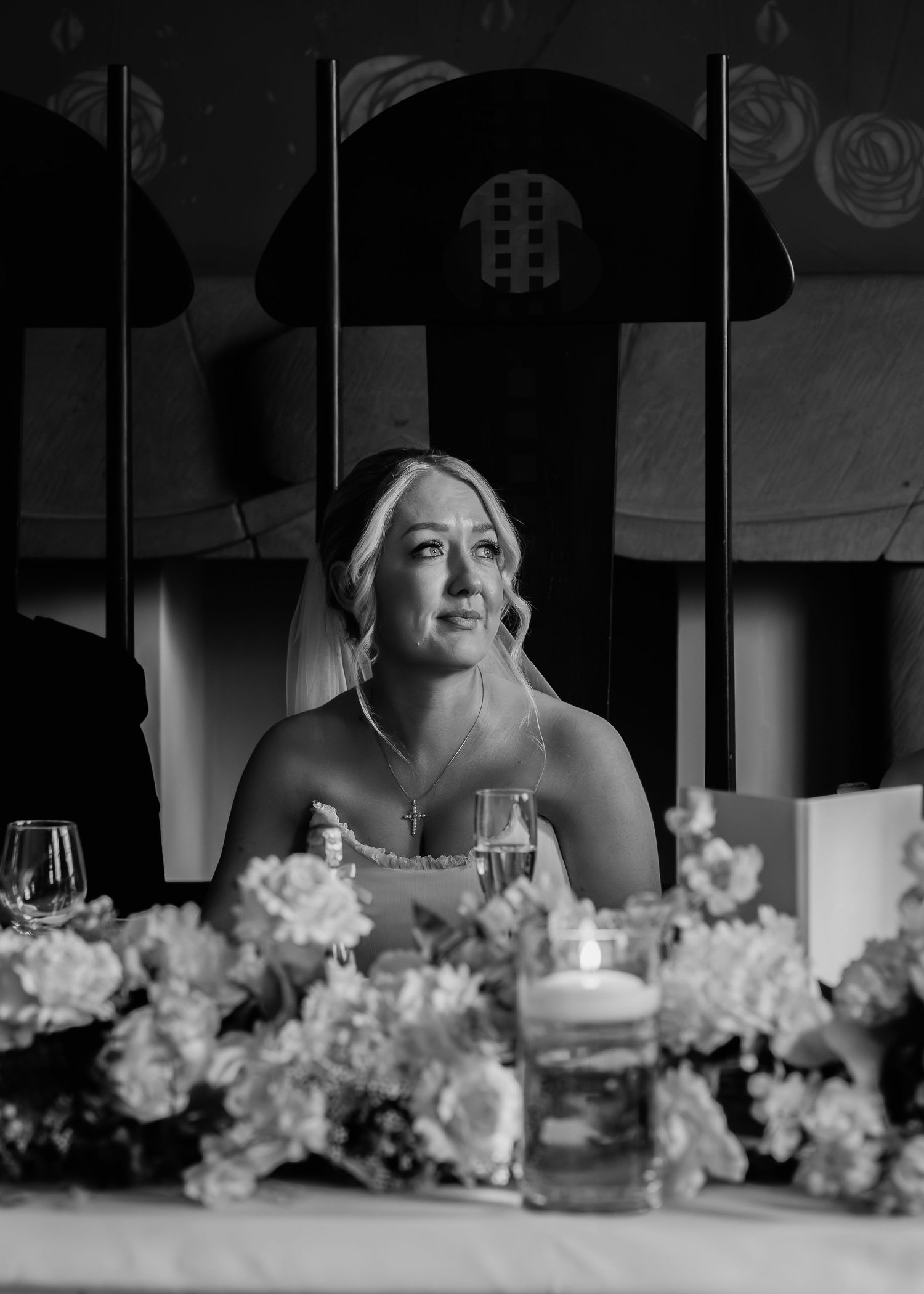 Black and white photo of a bride seated at a floral-decorated reception table, gazing thoughtfully in elegant wedding attire.