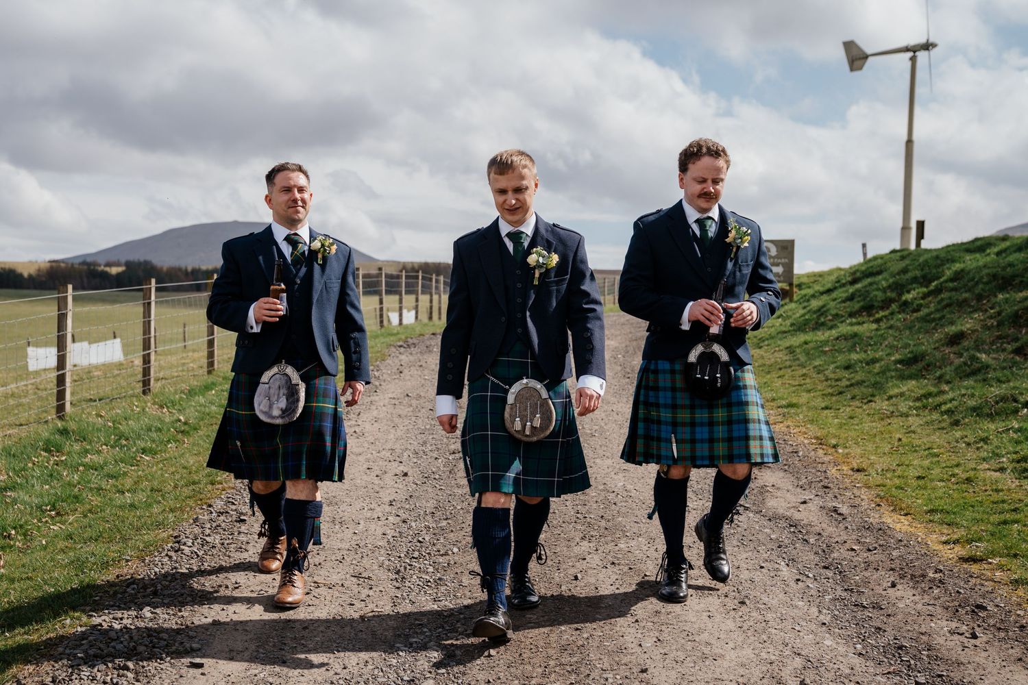 Three groomsmen in traditional Scottish kilts walking along a gravel path at an outdoor wedding venue.