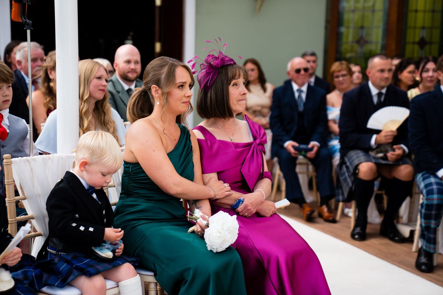 Guests seated at a wedding ceremony, women in elegant green and purple dresses, with a young child in traditional Scottish attire.