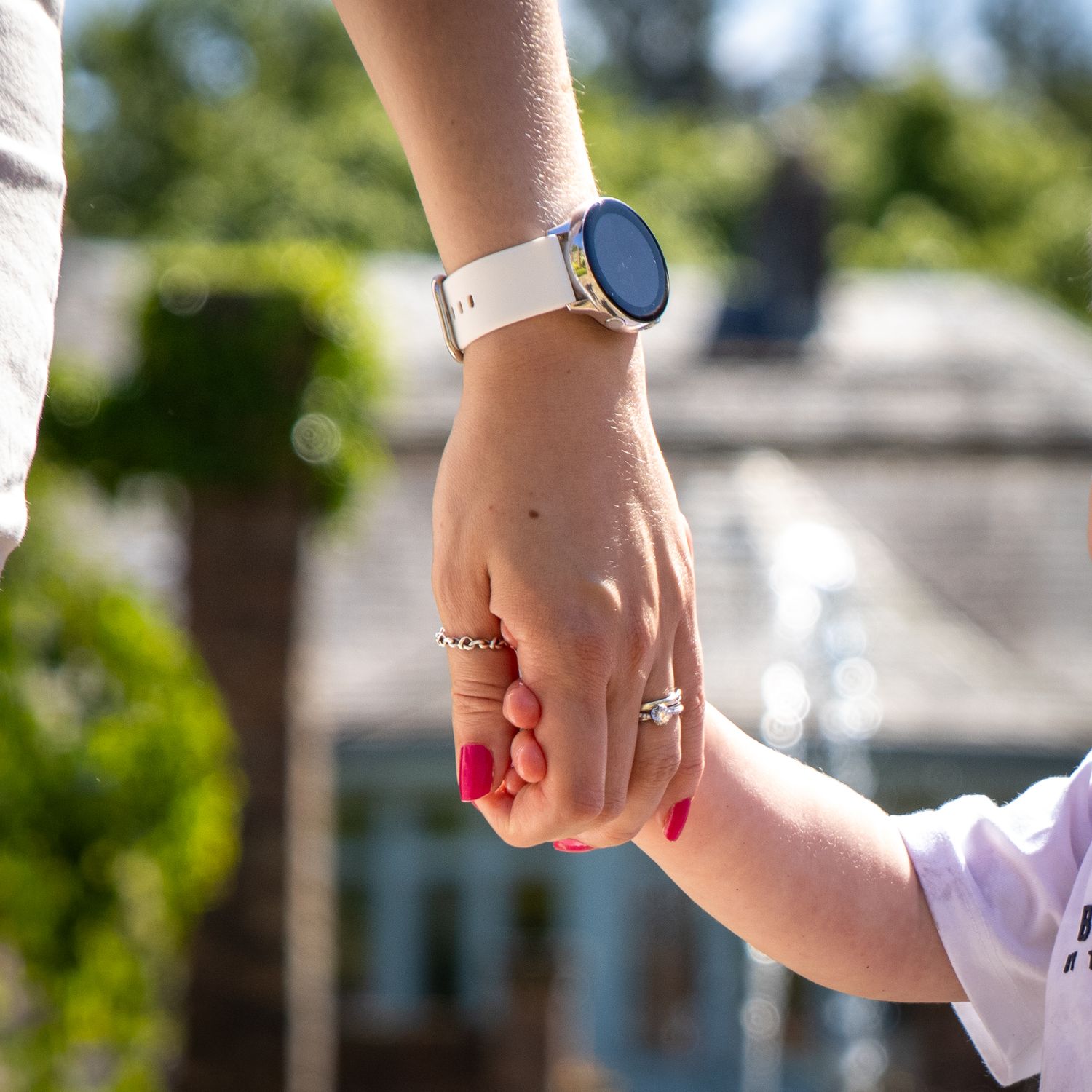 Close-up of hands with red nail polish and jewelry against a sunny outdoor background.