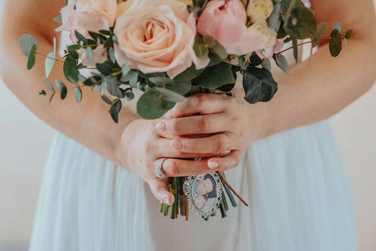 Bride holding a blush pink rose and eucalyptus wedding bouquet with a locket charm, wearing a light blue gown.