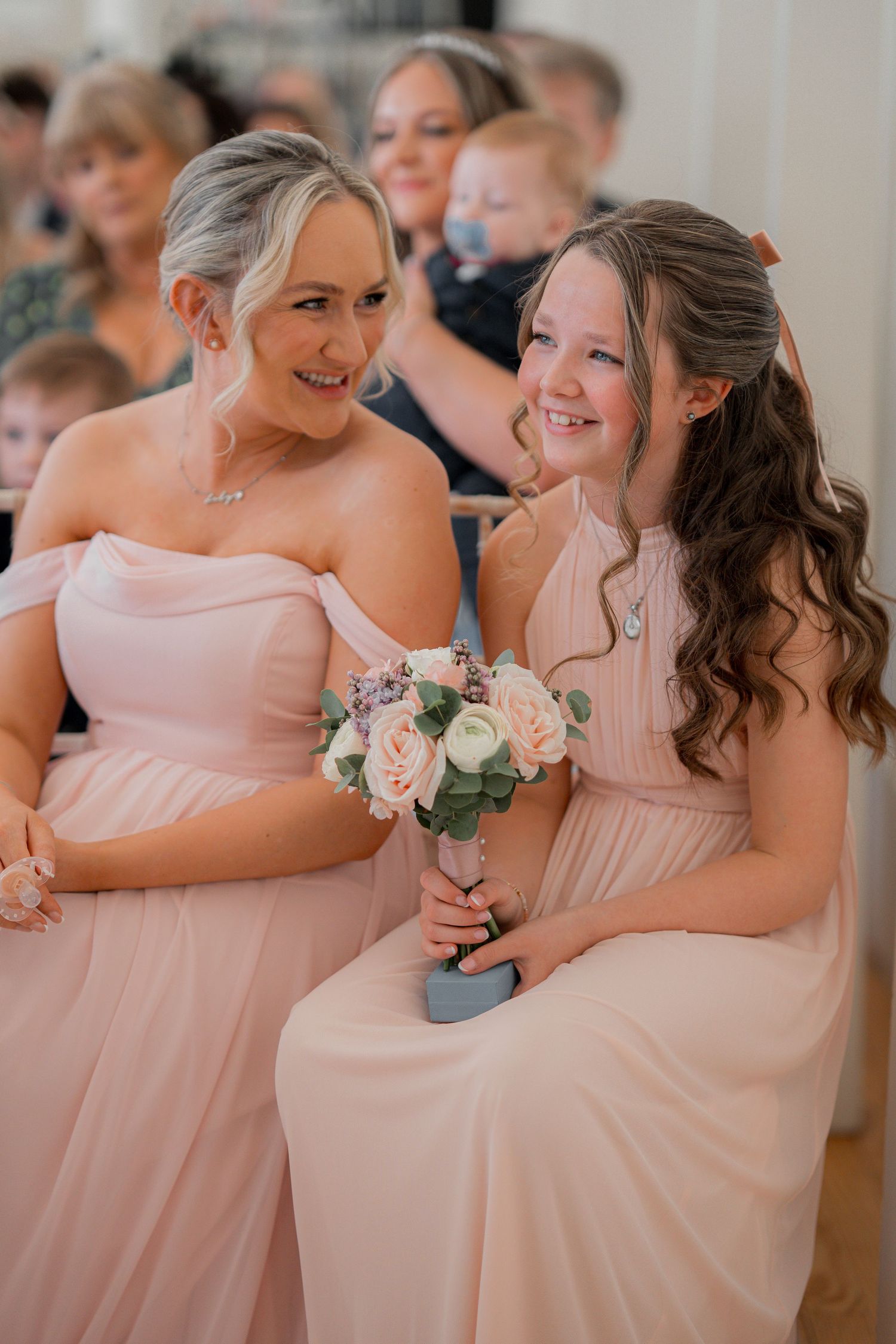 Two bridesmaids in blush pink dresses smile joyfully at a wedding ceremony, the younger holding a small bouquet of roses.