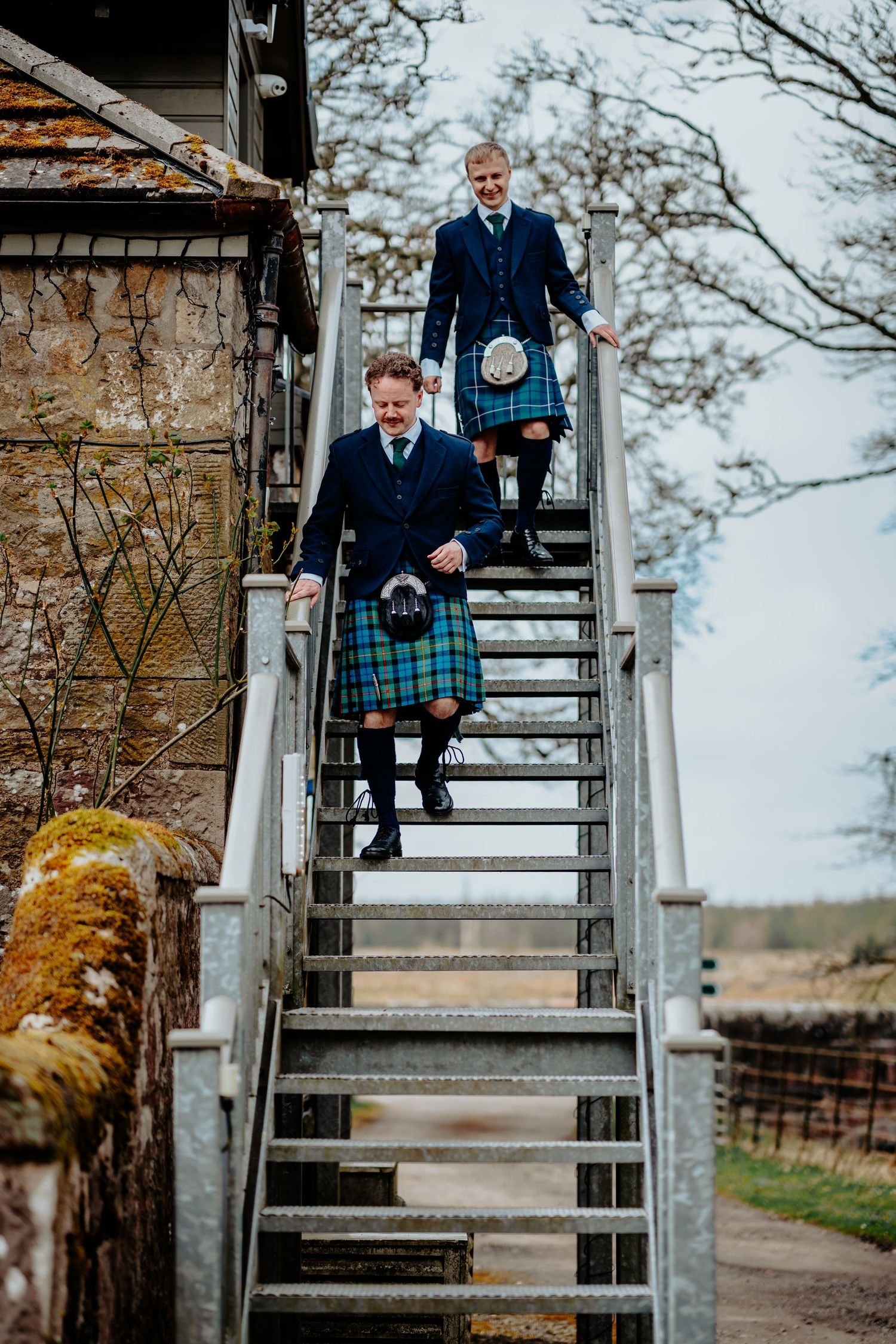 Two men in Scottish kilts and navy jackets descend outdoor metal stairs at a historic stone venue.