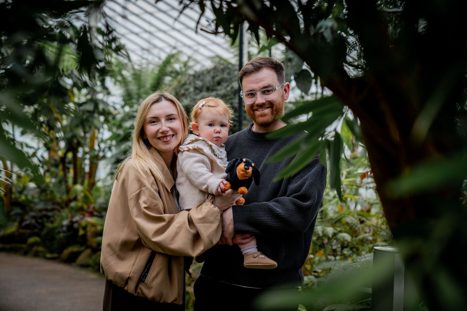 A smiling family with a baby poses among lush tropical plants inside a greenhouse botanical garden.