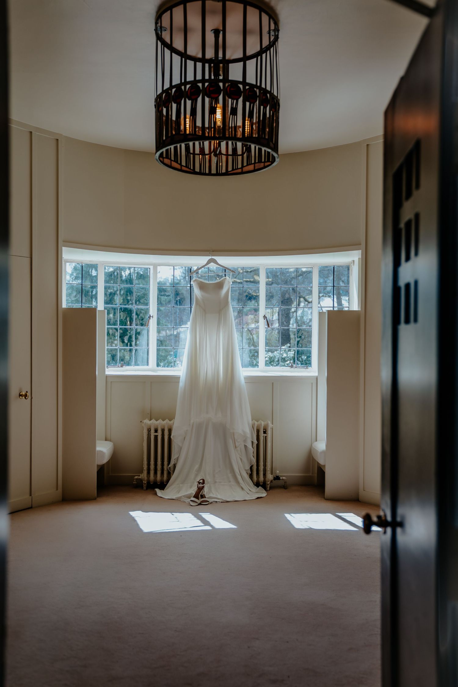 A elegant white wedding dress hanging in a sunlit bay window room with a decorative chandelier overhead.