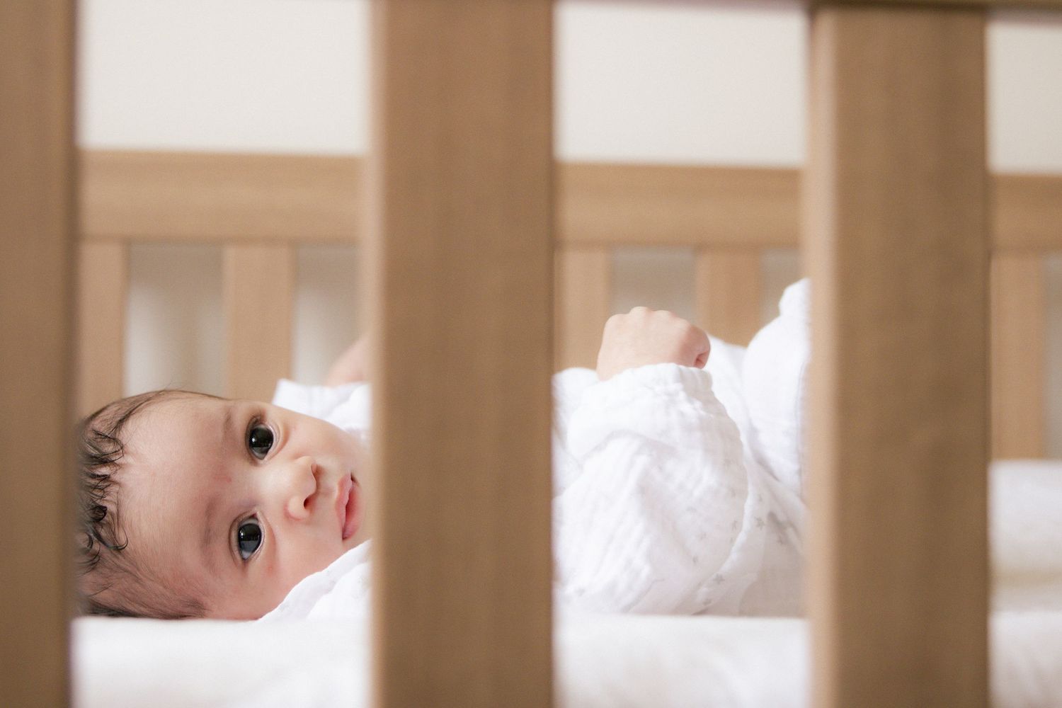 Newborn baby peeking through wooden crib slats in natural light.