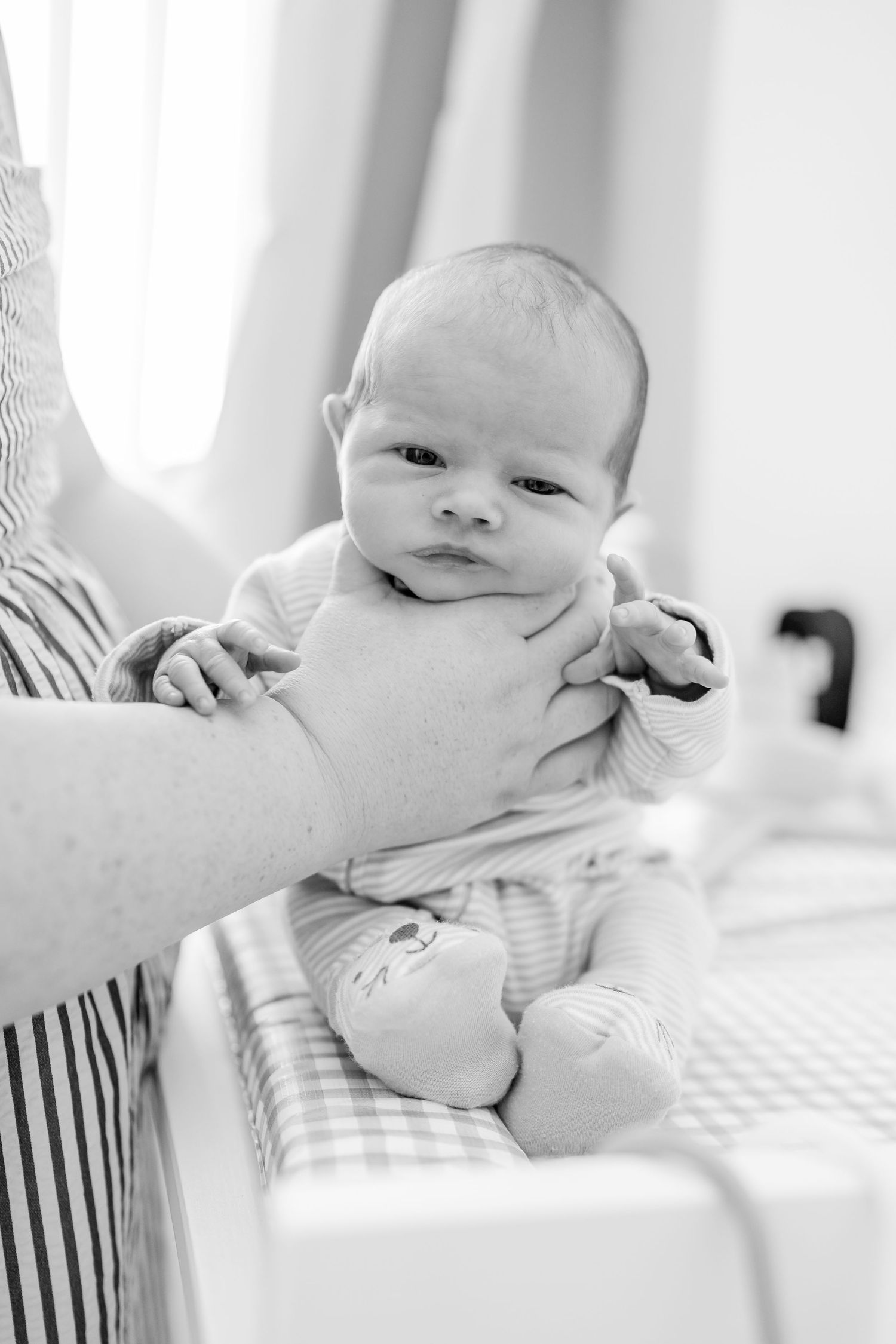 Black and white photo of a newborn baby being held upright by an adult's hands near a crib.
