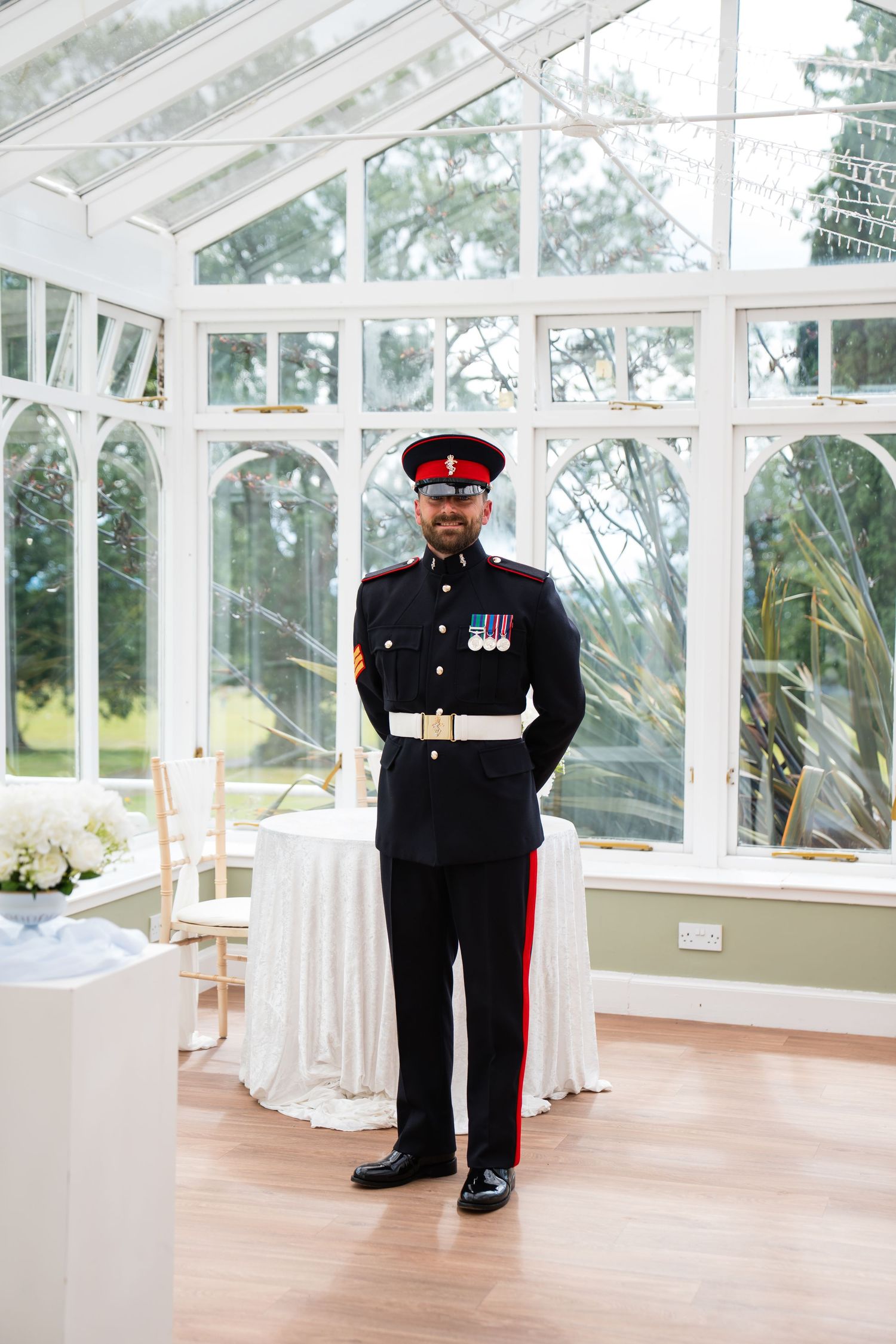 British military officer in ceremonial uniform with medals stands in an elegant glass conservatory wedding venue.