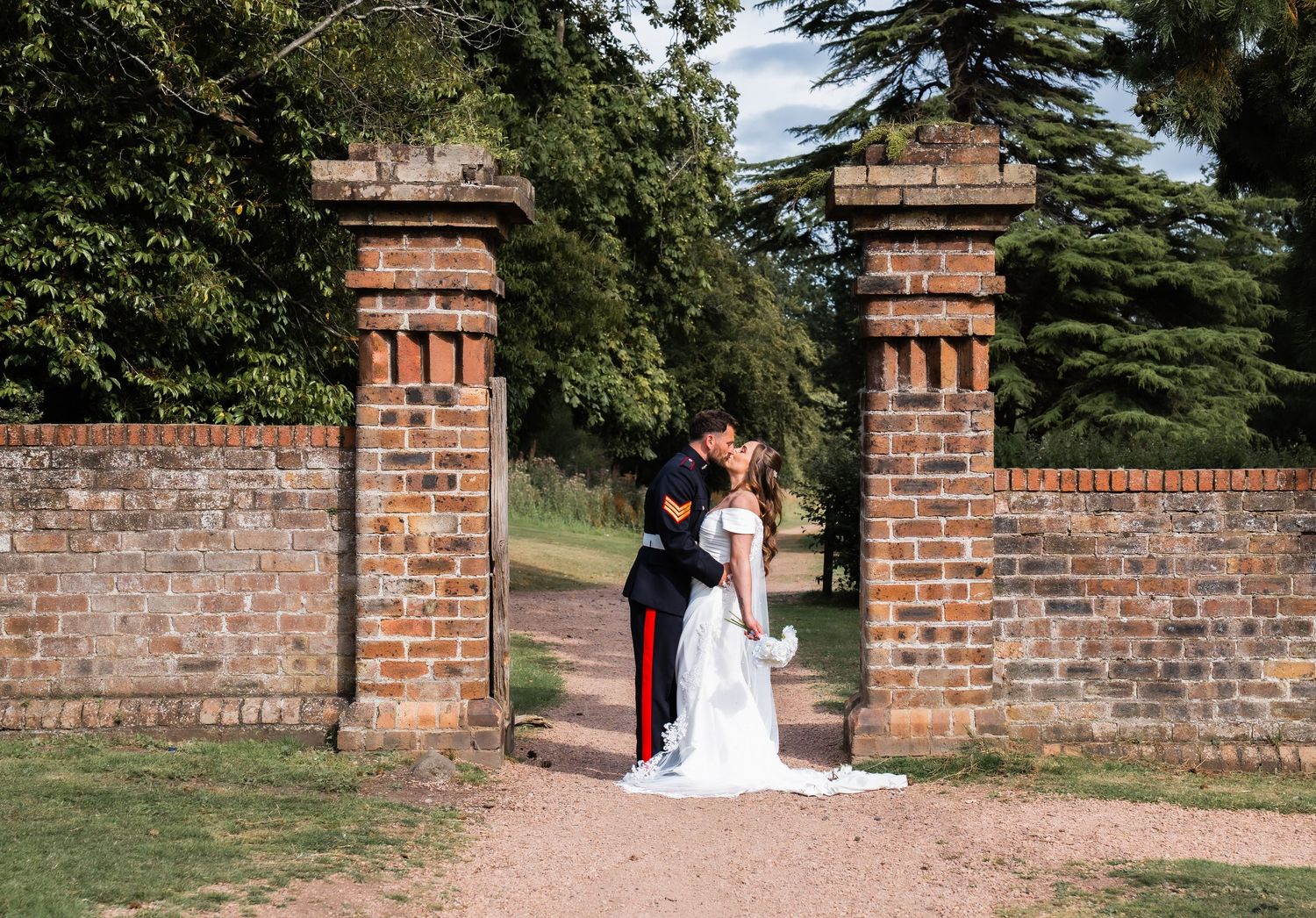 Bride and groom kissing in a brick gate entrance surrounded by lush green trees at an elegant outdoor wedding venue.