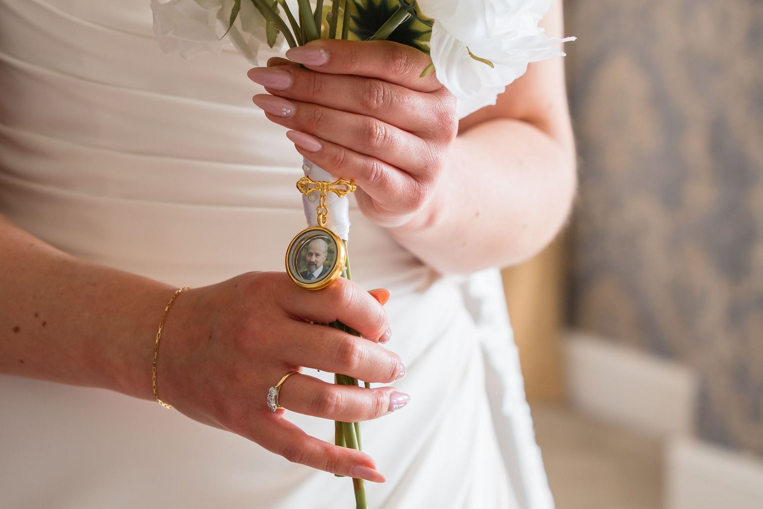 Bride holding white bouquet with gold memorial locket charm, wearing white dress and delicate bracelet.