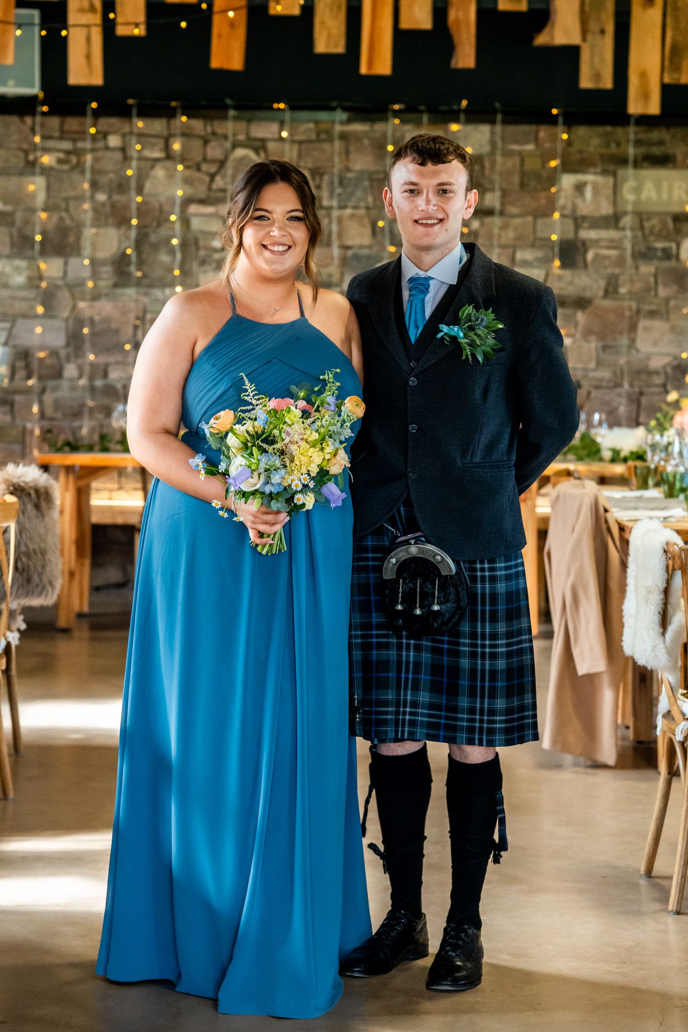 Bridesmaid in blue gown holding floral bouquet stands with groomsman in Scottish tartan kilt at wedding reception.