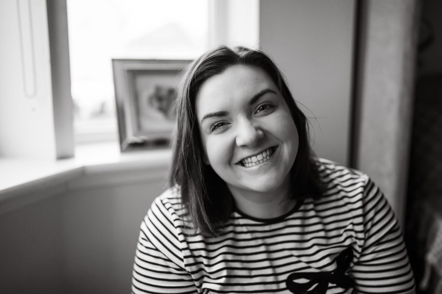 Black and white portrait of a smiling young woman in a striped shirt, sitting near a window indoors.