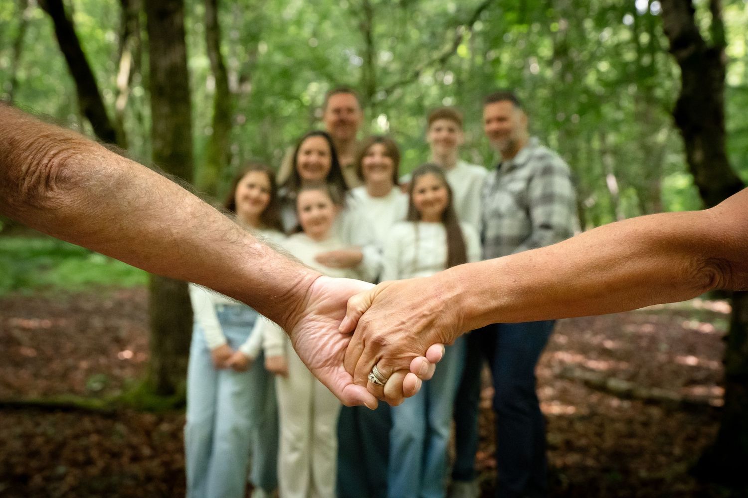 Elderly couple holding hands in forest with multi-generational family smiling in background.