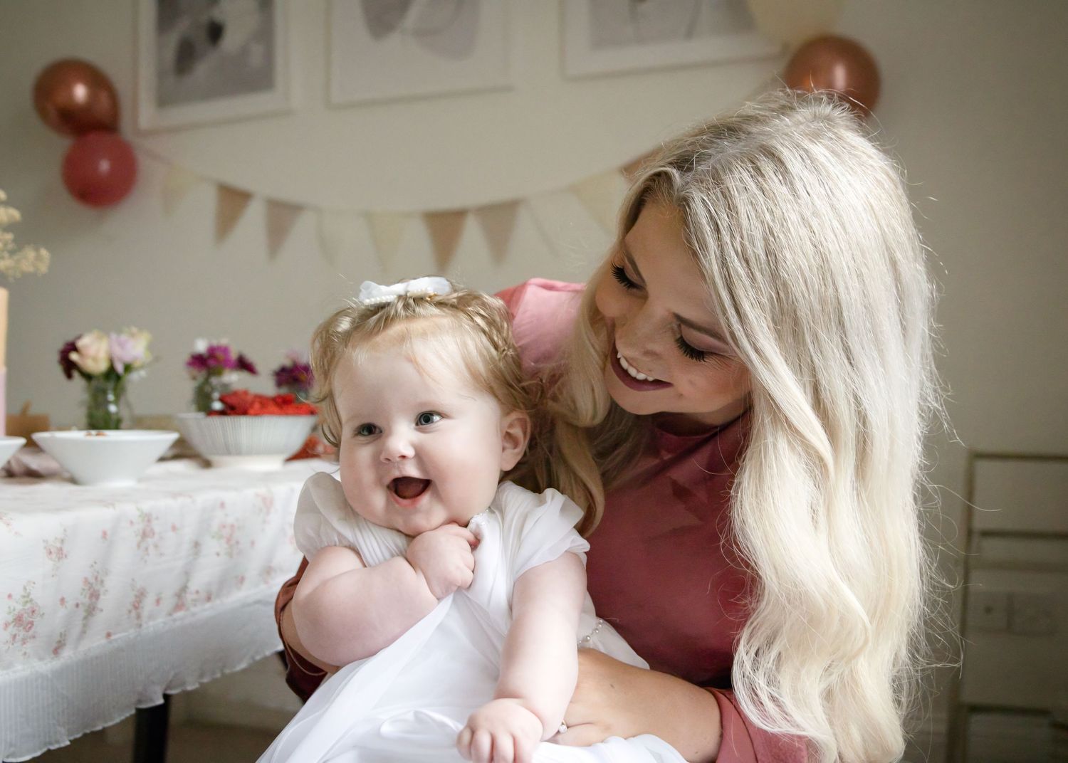 A smiling blonde woman holds a happy baby girl in a white dress at a first birthday party celebration.