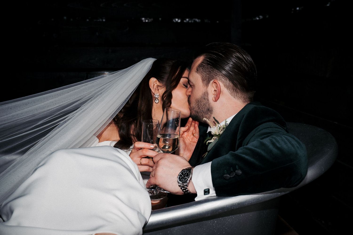 Bride and groom sharing a kiss while holding champagne glasses in a dark romantic setting on their wedding day.