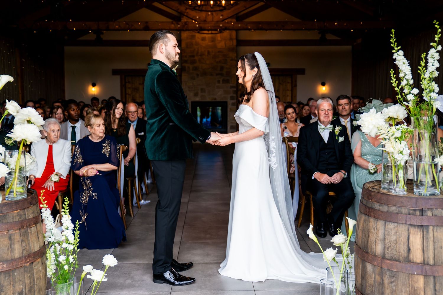 Bride and groom hold hands at rustic barn wedding ceremony aisle lined with white floral arrangements and barrel decor.