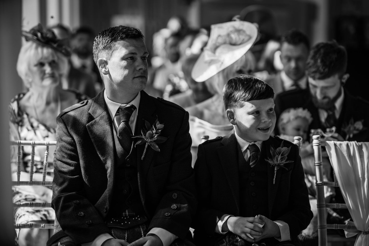 Two boys in suits with boutonnieres sit attentively during a wedding ceremony in black and white photo.