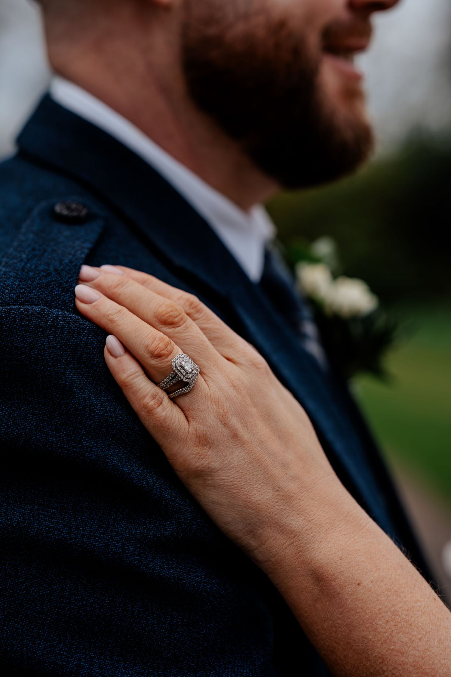 Close-up of bride's hand with diamond wedding ring resting on groom's navy blue tweed suit jacket.