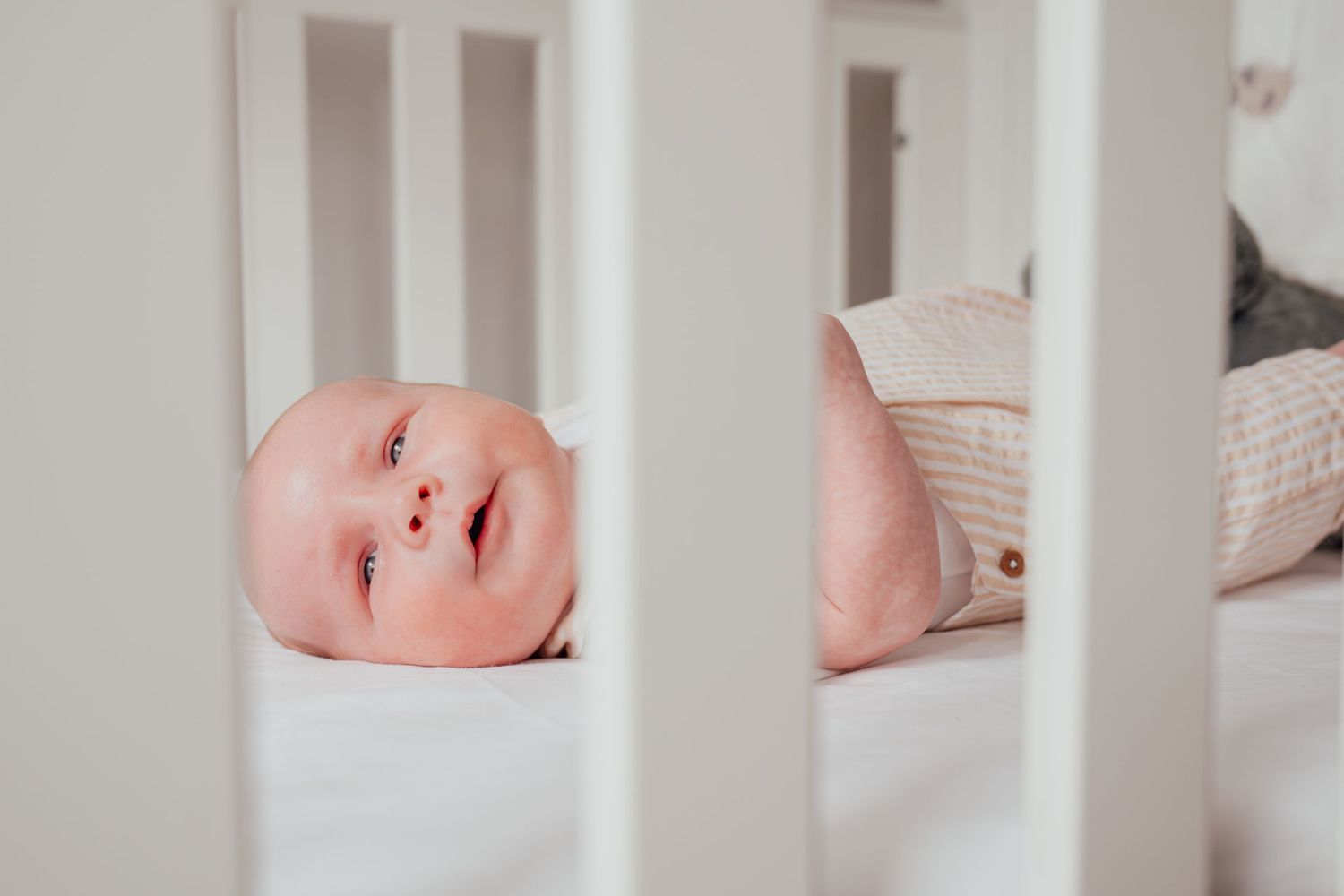 Smiling baby lying awake in a white wooden crib, wearing a striped onesie on a white sheet.