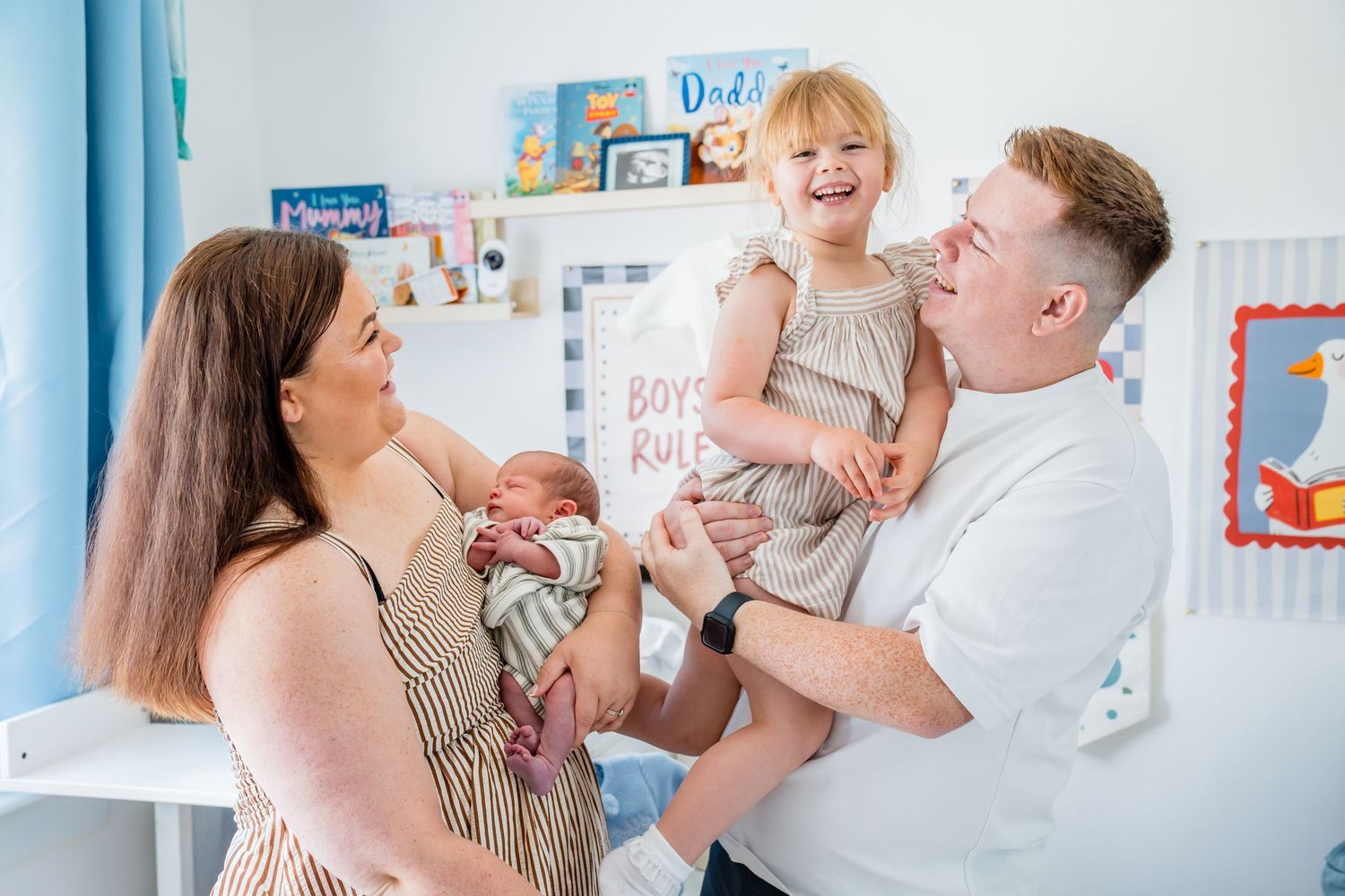 A happy family of four in a nursery, parents holding a newborn and laughing toddler girl in a bright decorated room.