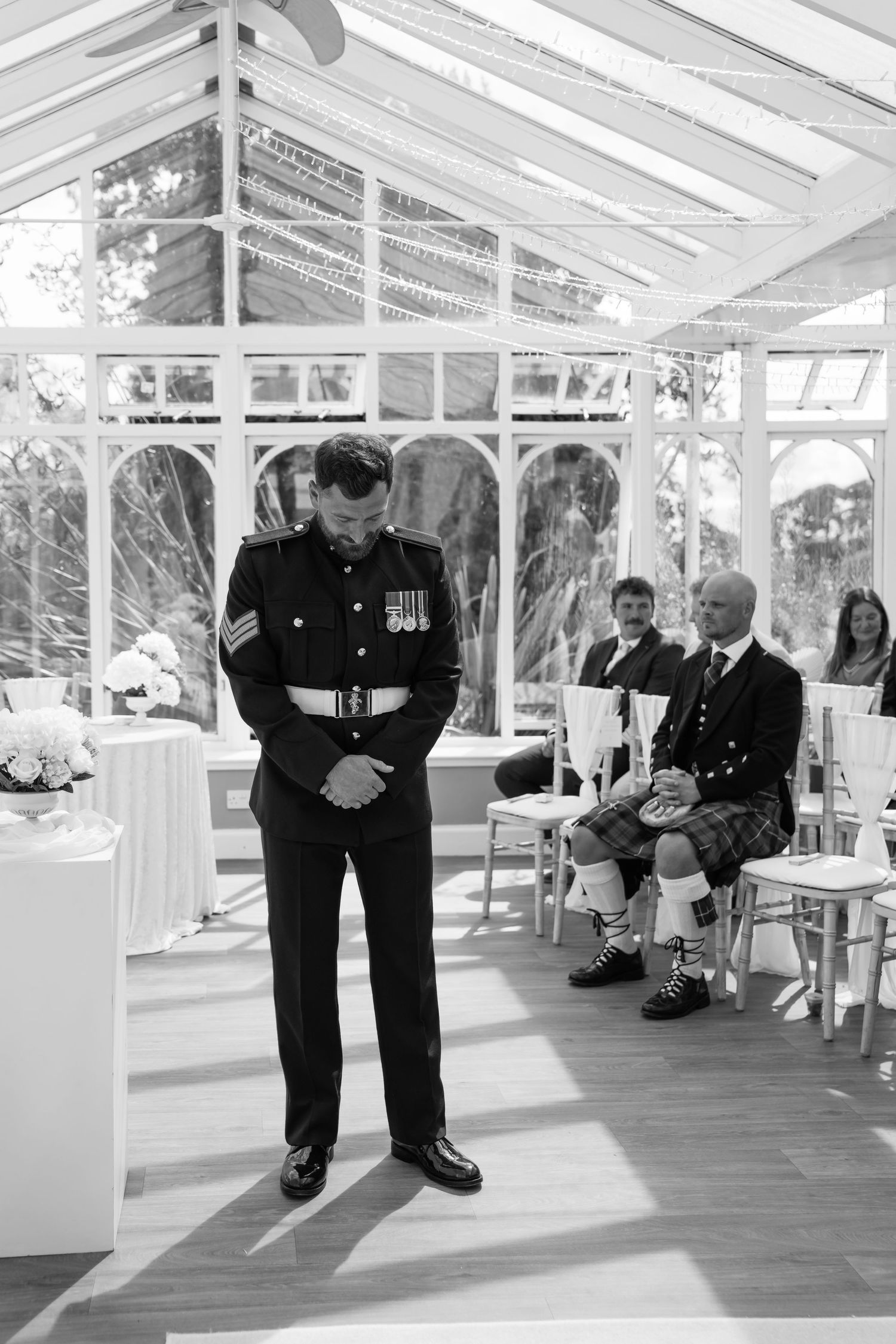Groom in military uniform bows head at wedding altar in elegant glass conservatory venue, black and white photo.
