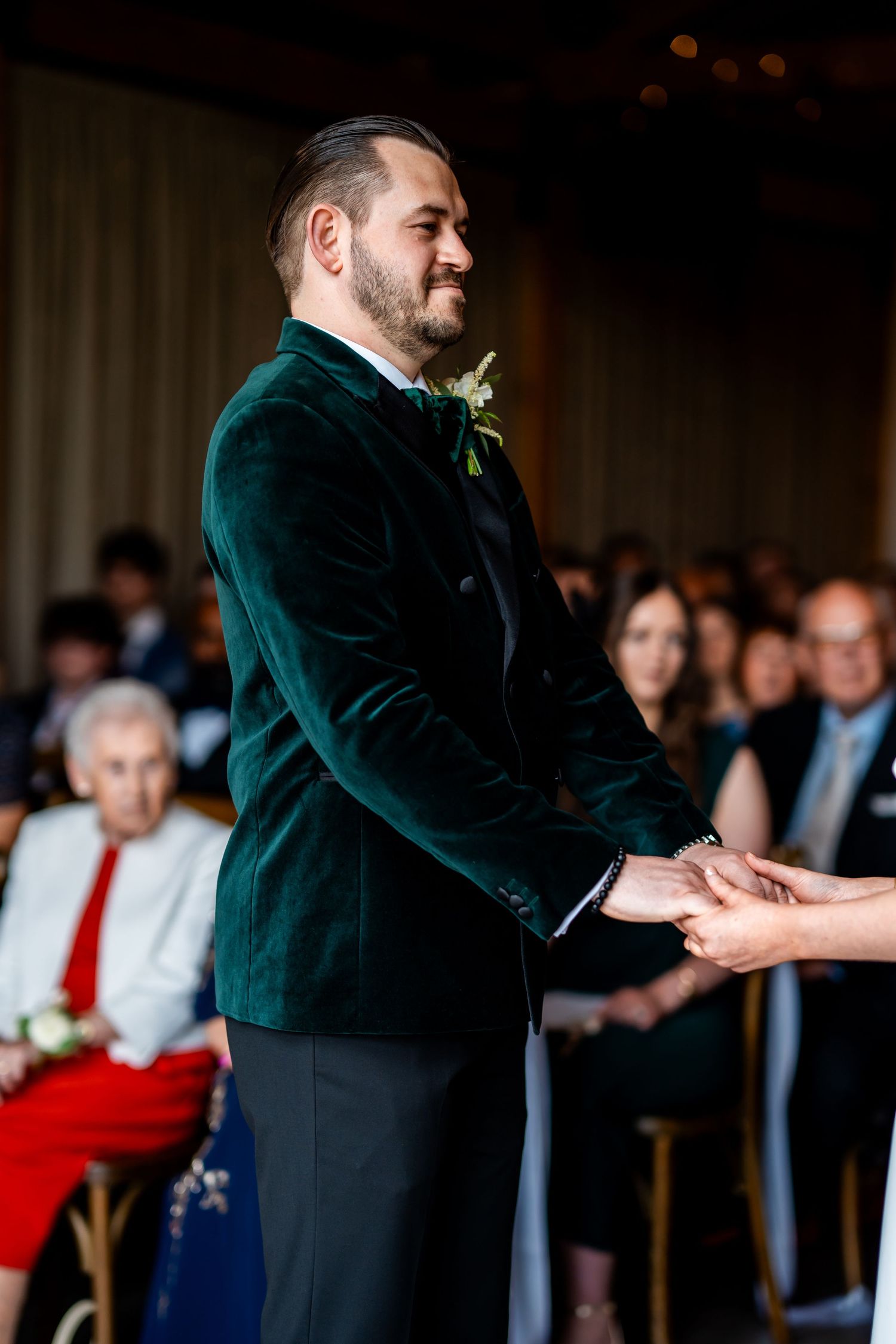 Groom in green velvet tuxedo jacket smiling while holding hands with bride during indoor wedding ceremony.
