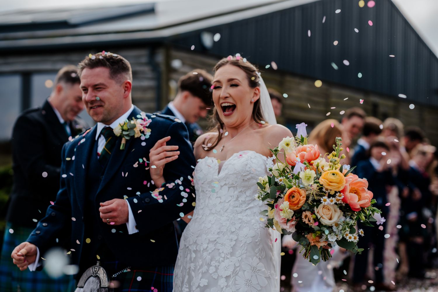 Joyful bride and groom walk through colorful confetti shower, bride holding vibrant bouquet at outdoor wedding celebration.