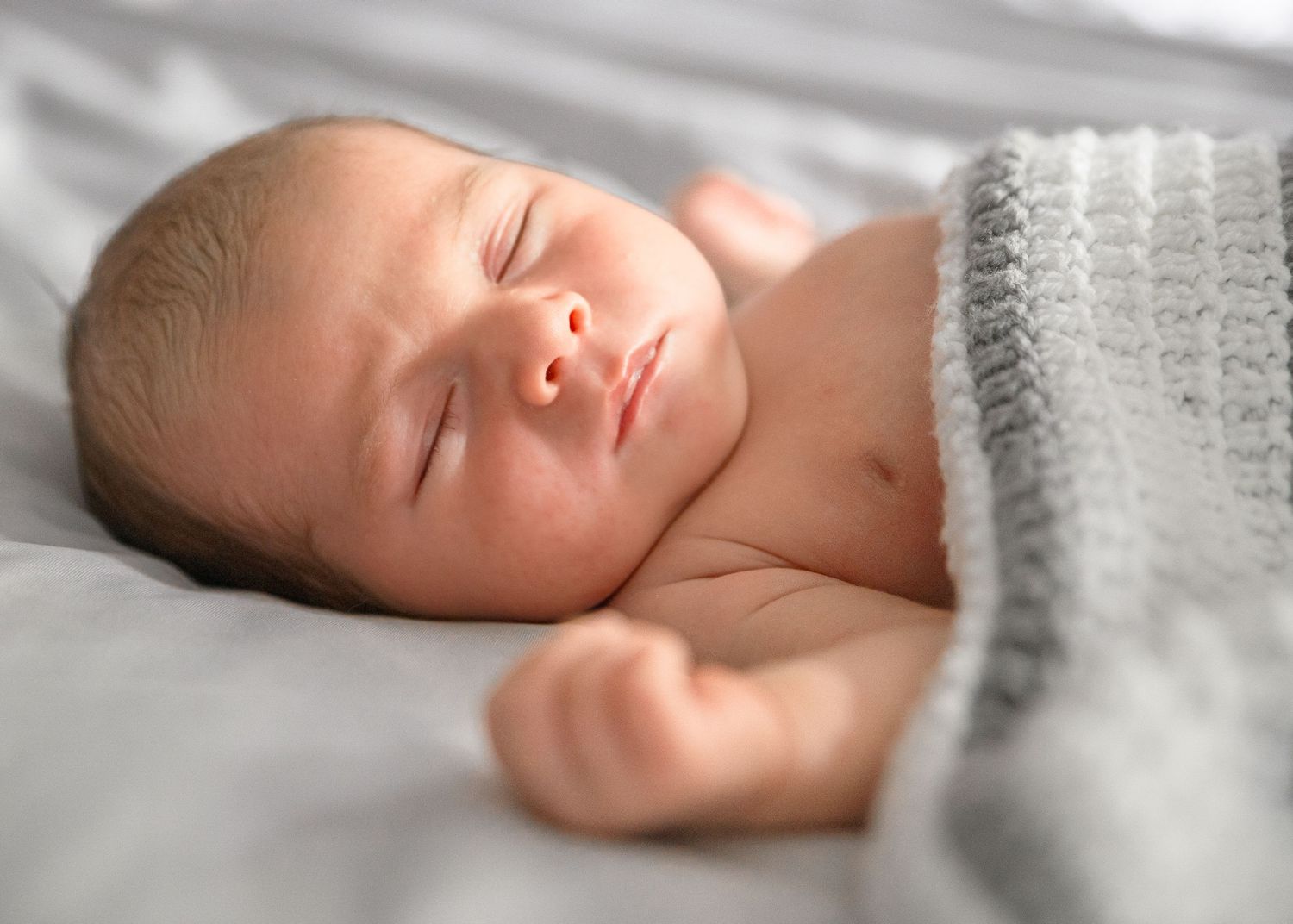 Peaceful newborn baby sleeping soundly wrapped in a soft gray knit blanket on white bedding.